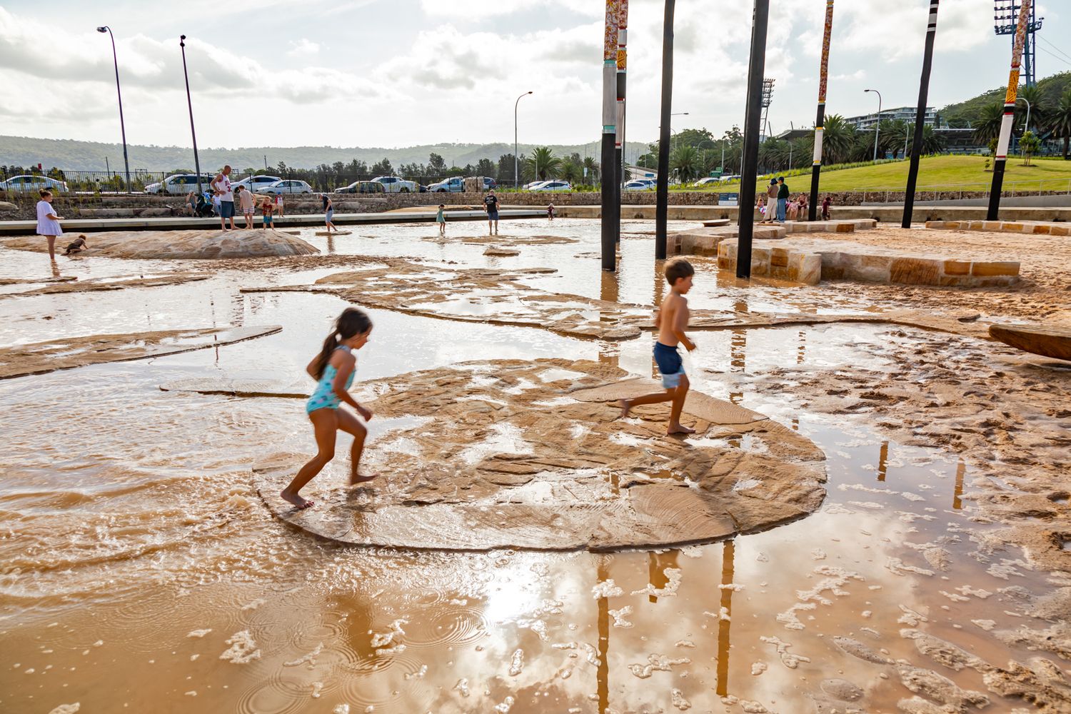 Two children play in shallow muddy water at a splash park with tall poles and hills in the background.