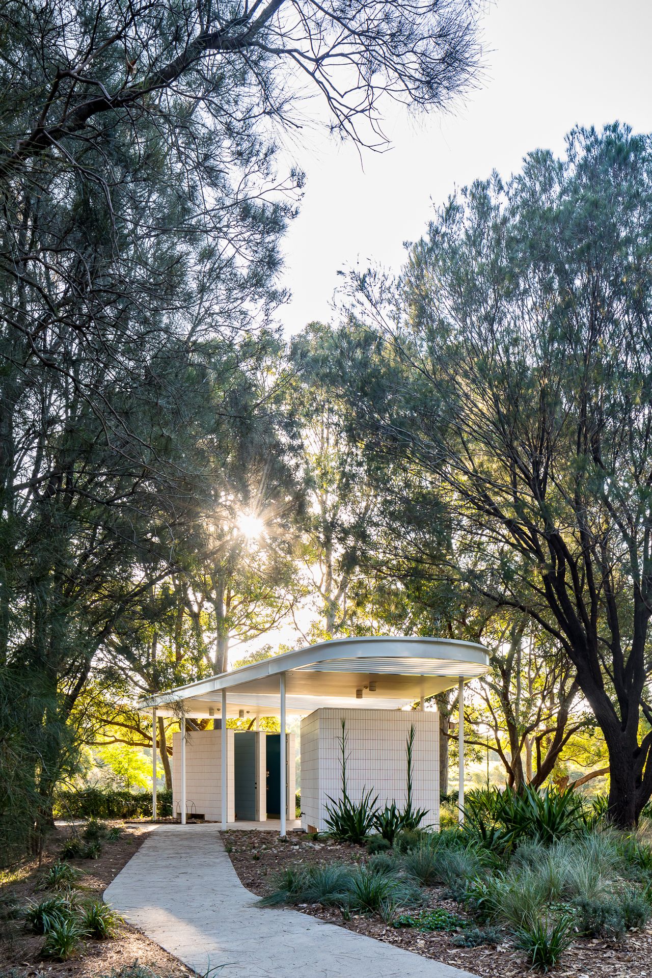 Sunlight filters through trees onto a small white pavilion with a curved roof, reached by a winding concrete path.