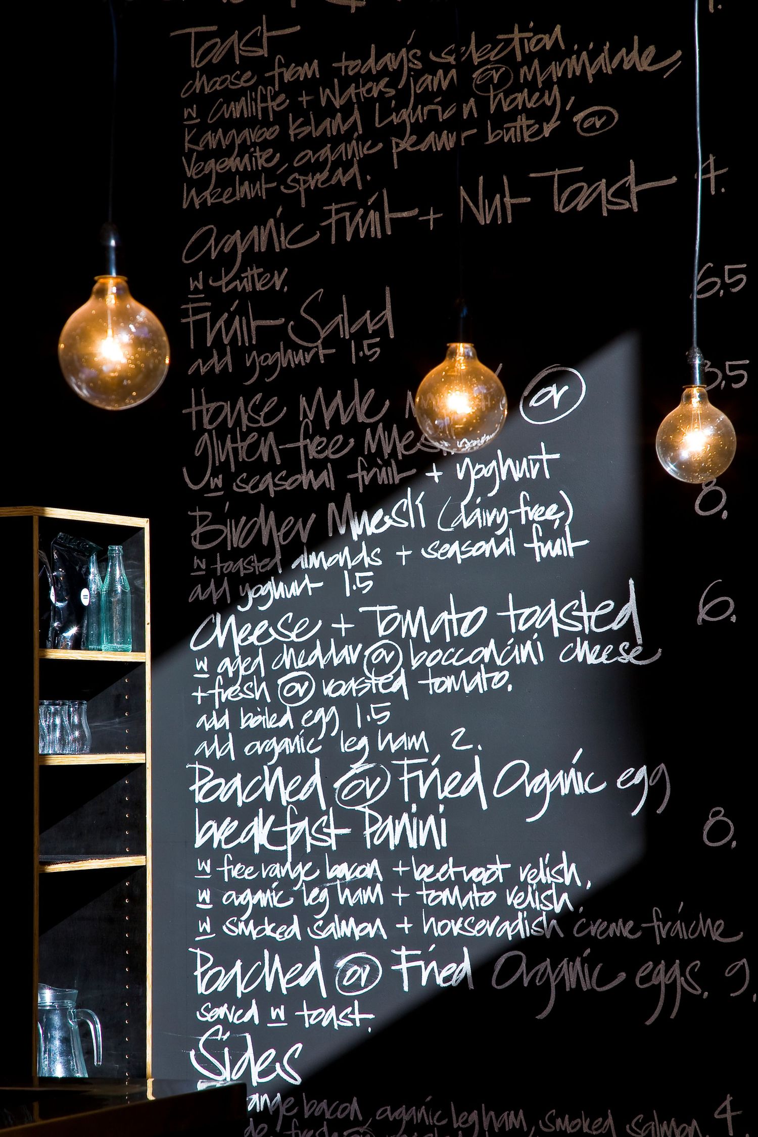 Chalkboard cafe menu handwritten in white chalk listing organic toasts, fruit salad, muesli and eggs, lit by glowing bulb pendant lights.