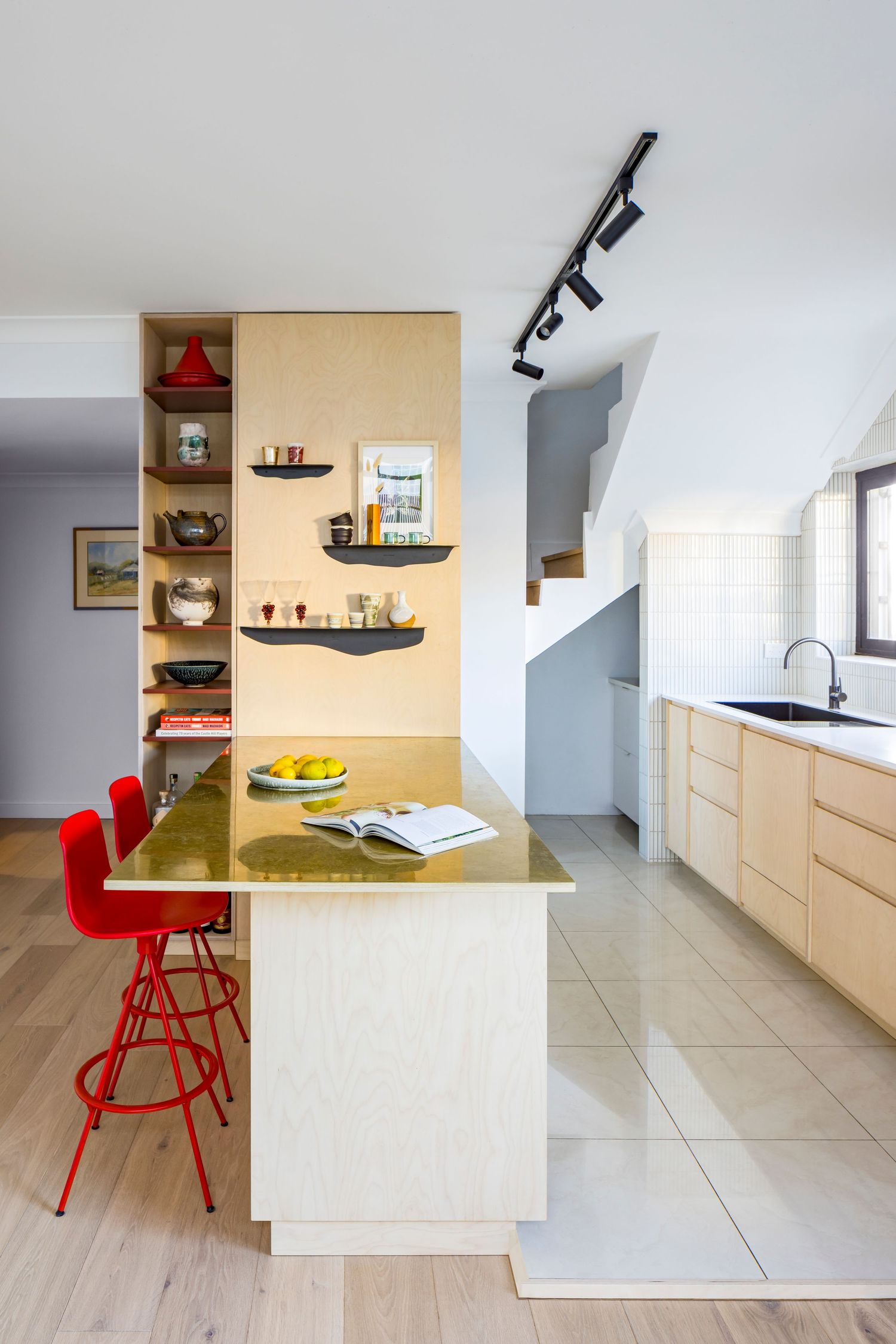 Modern kitchen with a plywood island, brass countertop, red stools, open shelving and black track lighting beside a staircase.