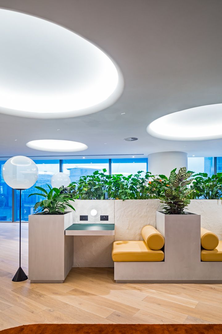 Modern office lounge with built-in planters, mustard yellow bench seating, a globe floor lamp, oval ceiling lights and city-view windows.