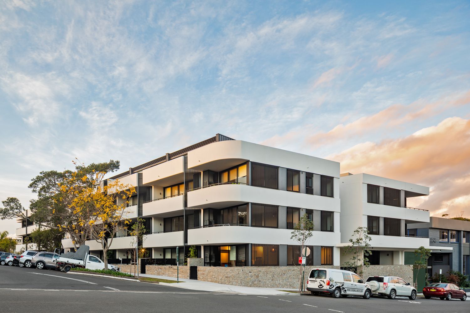 Modern white apartment building with curved balconies and large windows on a suburban street at sunset, with parked cars below.