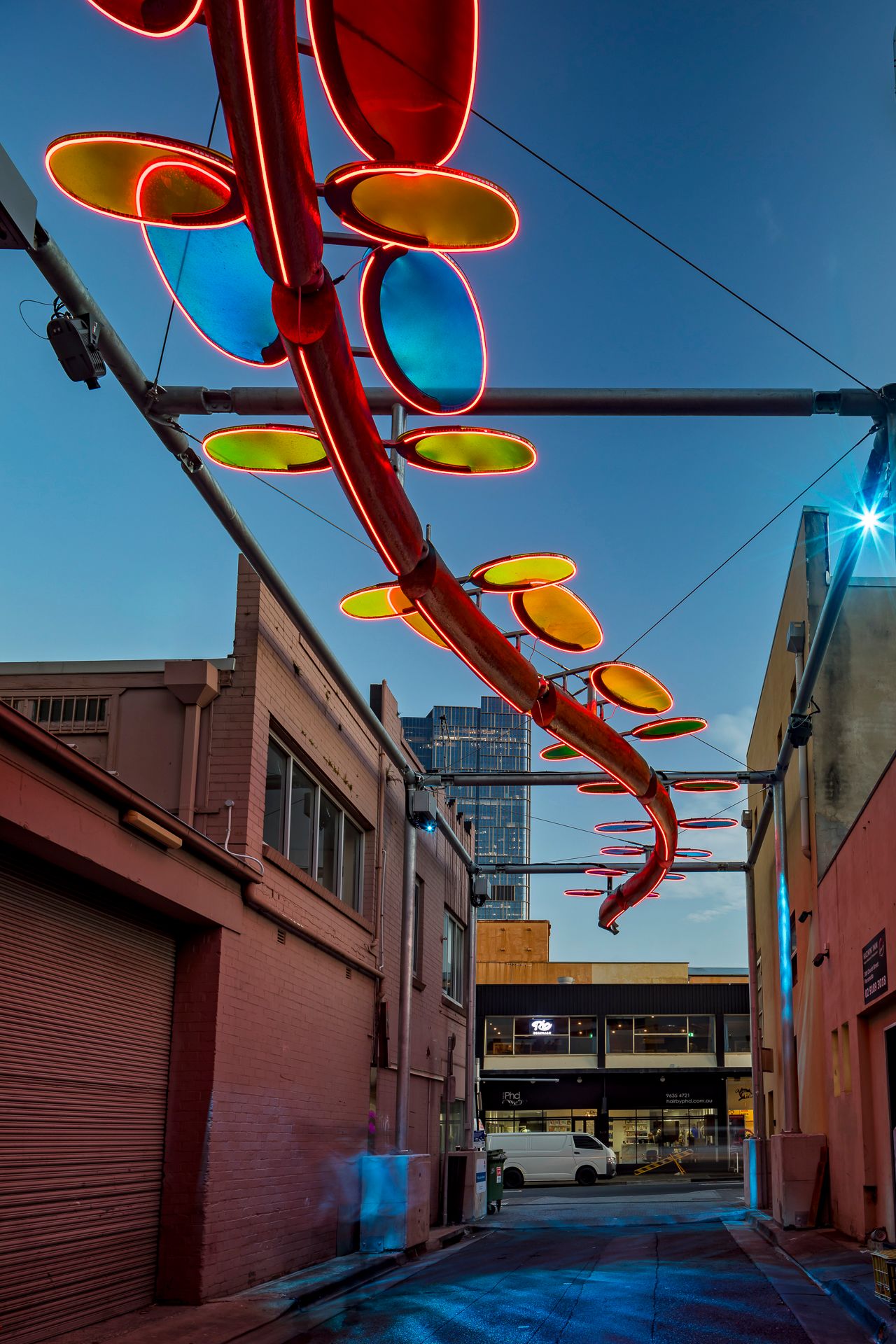 Red neon serpent sculpture with orange and blue oval discs suspended over a brick alley beneath a deep blue sky.