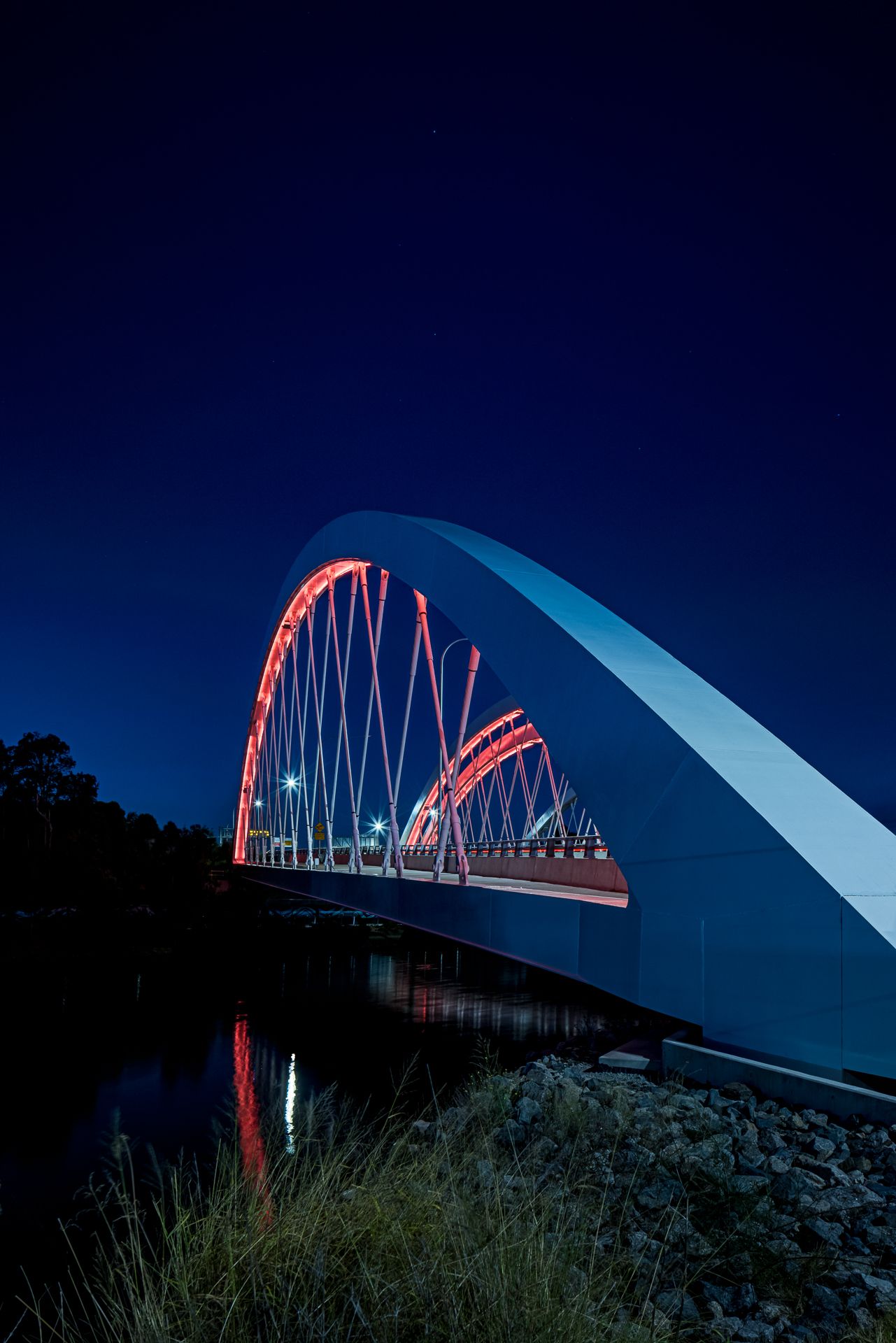 Large white bridge arch lit in red at night, with a rocky riverbank and grass in the foreground.