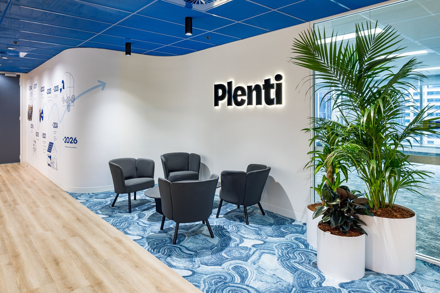 Plenti office reception with illuminated logo sign, black lounge chairs, potted palm plants and blue patterned carpet.