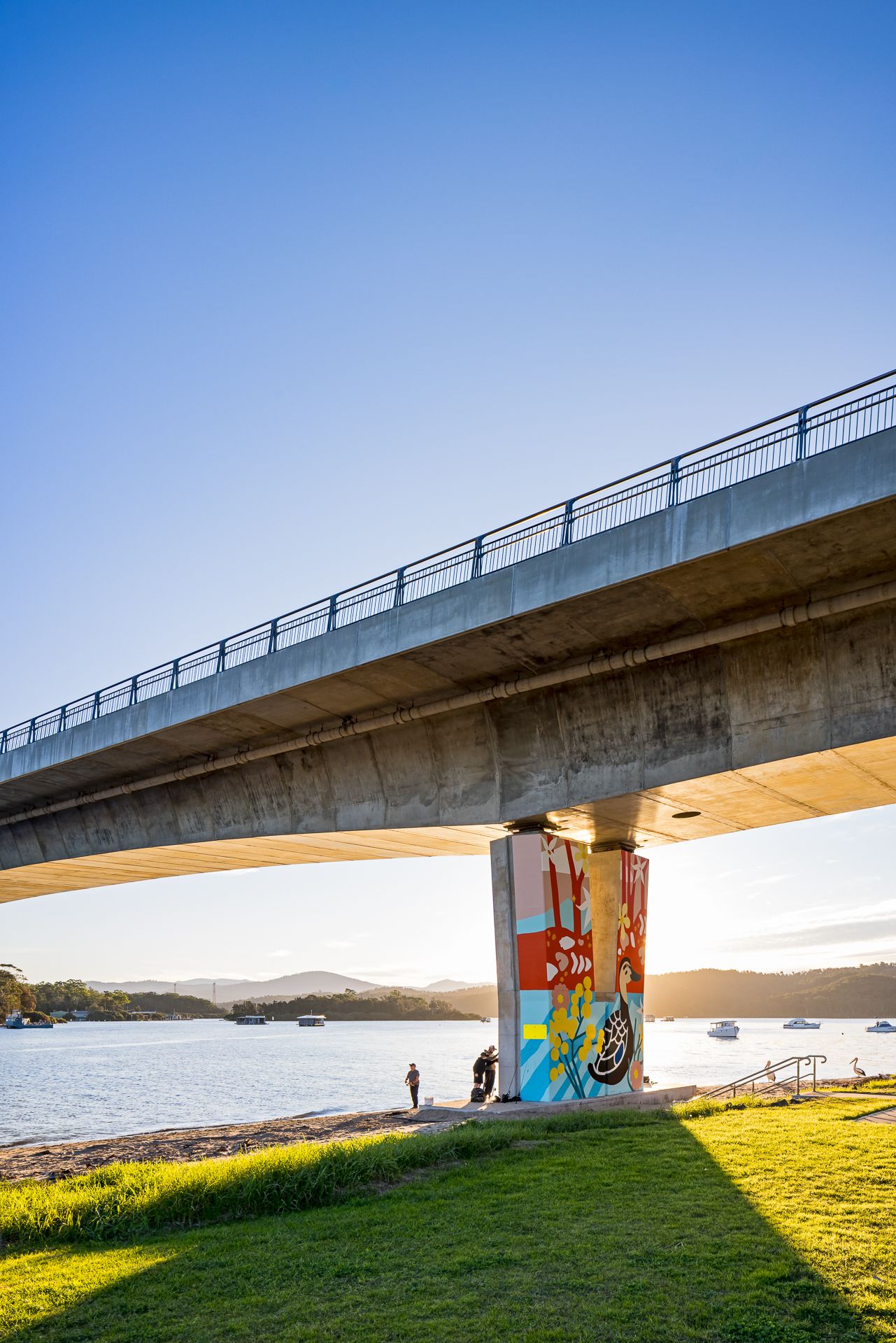Concrete bridge supported by a graffiti-style mural pillar above a grassy waterfront park, with boats on calm water at sunset.