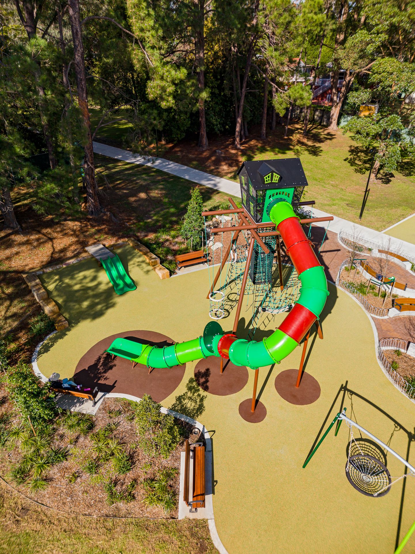 Overhead view of a playground with a long red and green tube slide, timber climbing tower, swings, benches and soft yellow rubber flooring.