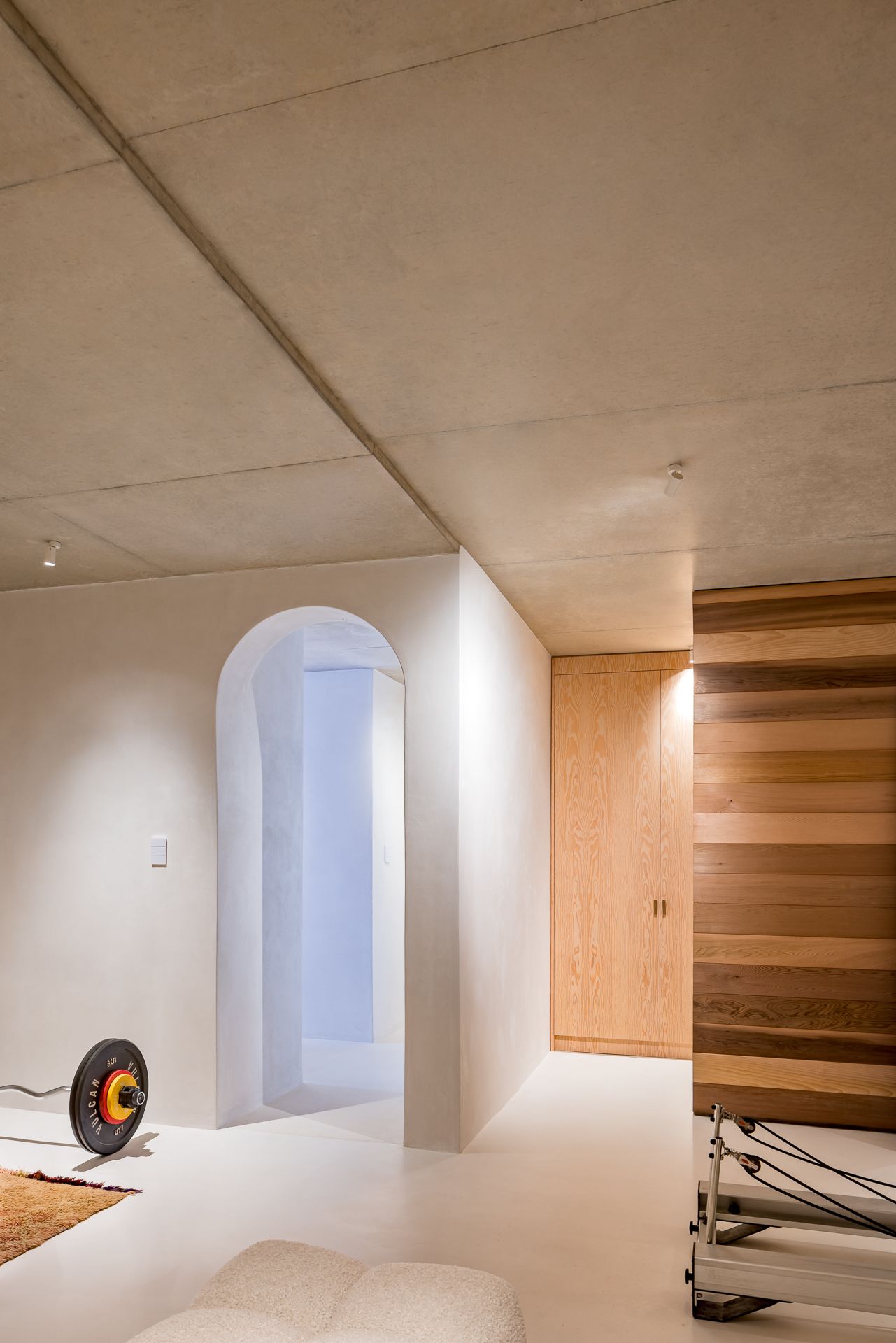 Modern interior with concrete ceiling, white arched passage, oak wood cabinetry, a bouclé ottoman and exercise equipment.