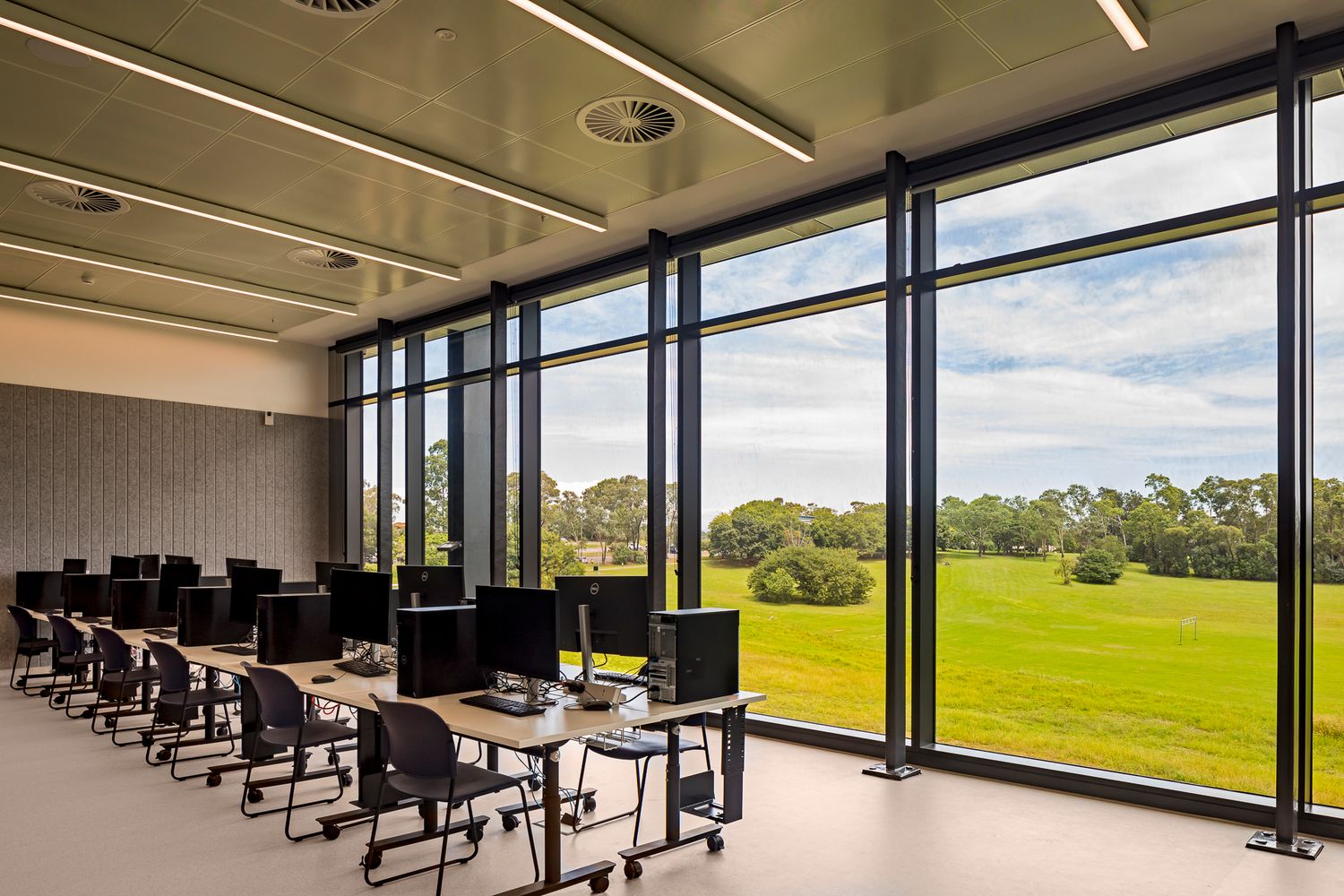 Bright computer classroom with rows of desktops and black chairs beside floor-to-ceiling windows overlooking a green lawn and trees.