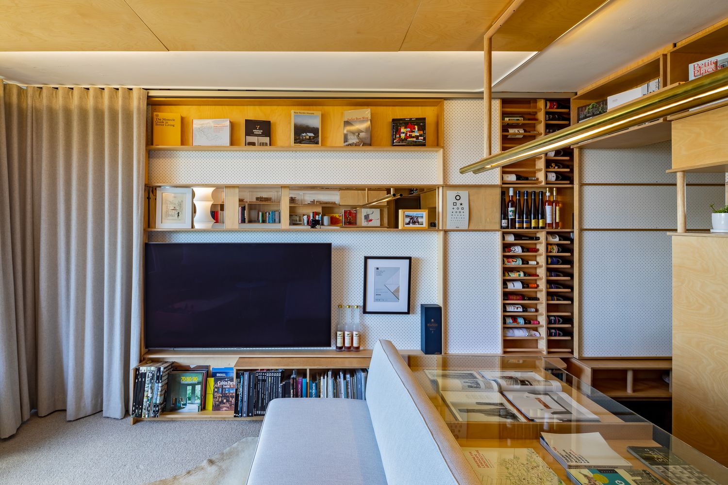 Apartment living area with a wall-mounted television, timber bookshelves, a sofa, and a beige curtain beside a glass-topped table.
