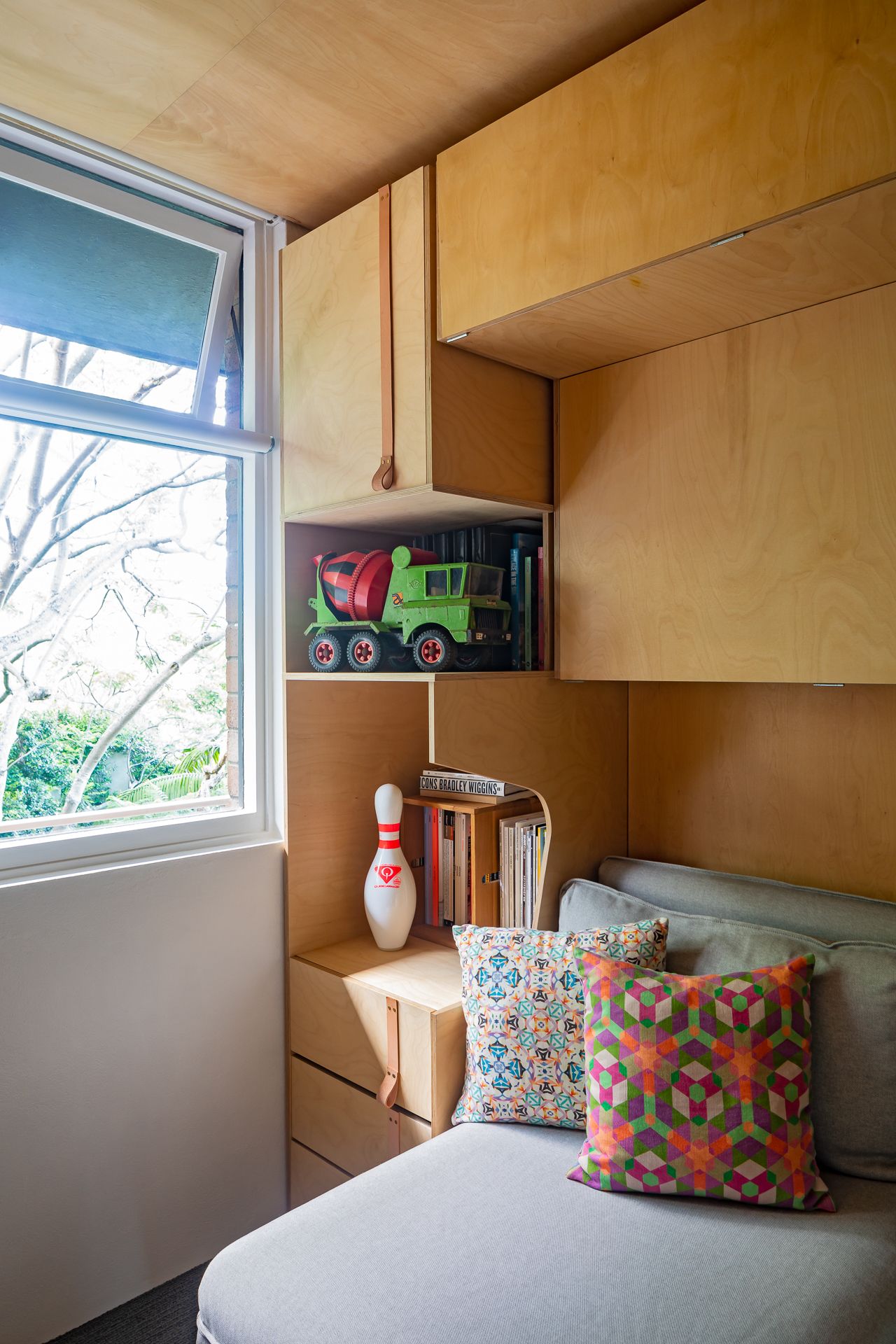 Corner of a plywood-lined room with a gray daybed, geometric cushions, a toy truck, books, a bowling pin and a window overlooking trees.
