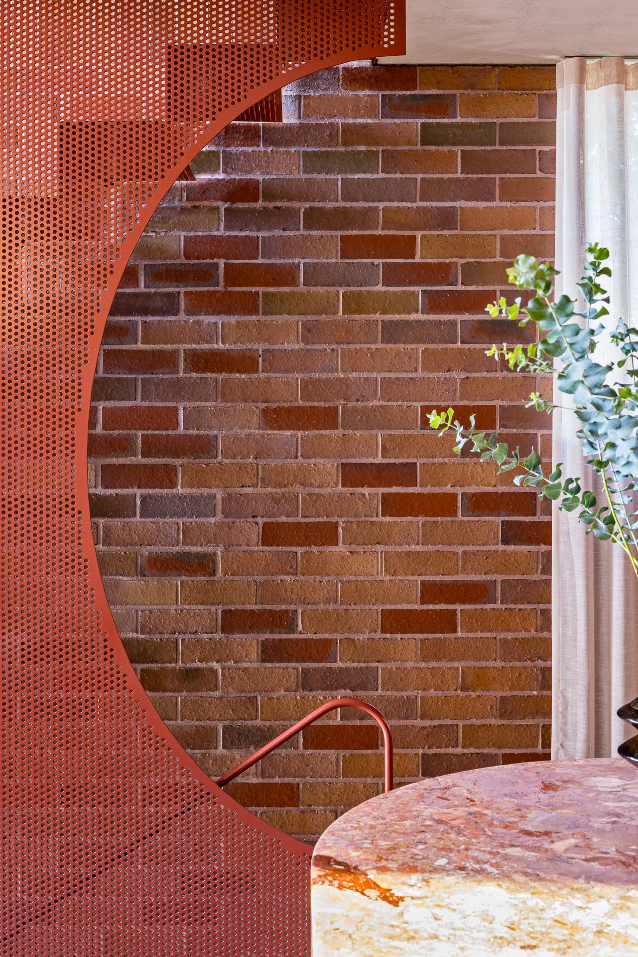 Red perforated metal screen with a circular cutout revealing a brick wall, a pink stone tabletop and eucalyptus leaves.