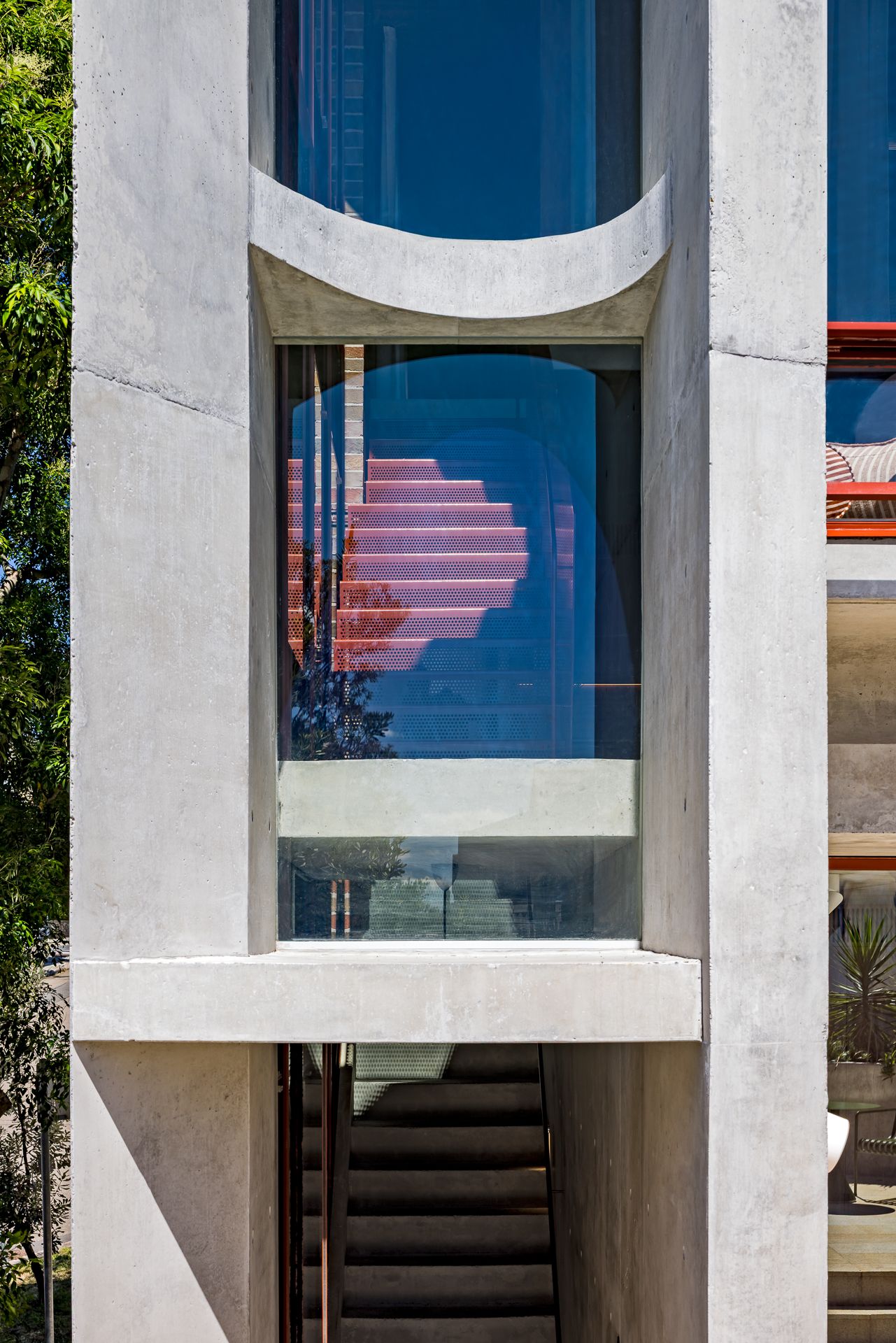 Close-up of a concrete window frame with a curved lintel reflecting red stairs and blue sky above an outdoor staircase.