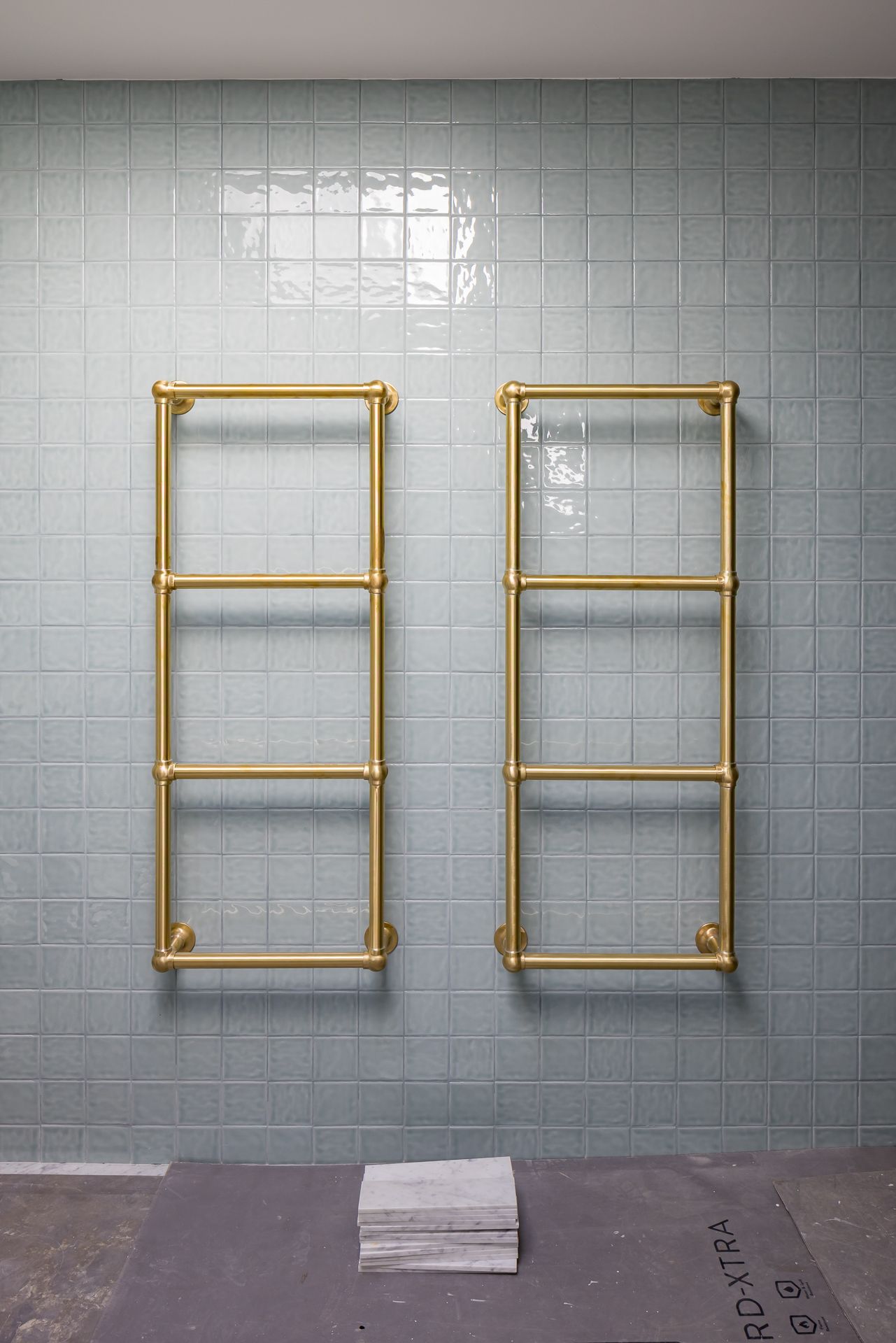 Two brass ladder-style heated towel rails mounted on a sage green glazed tile wall in an unfinished bathroom.