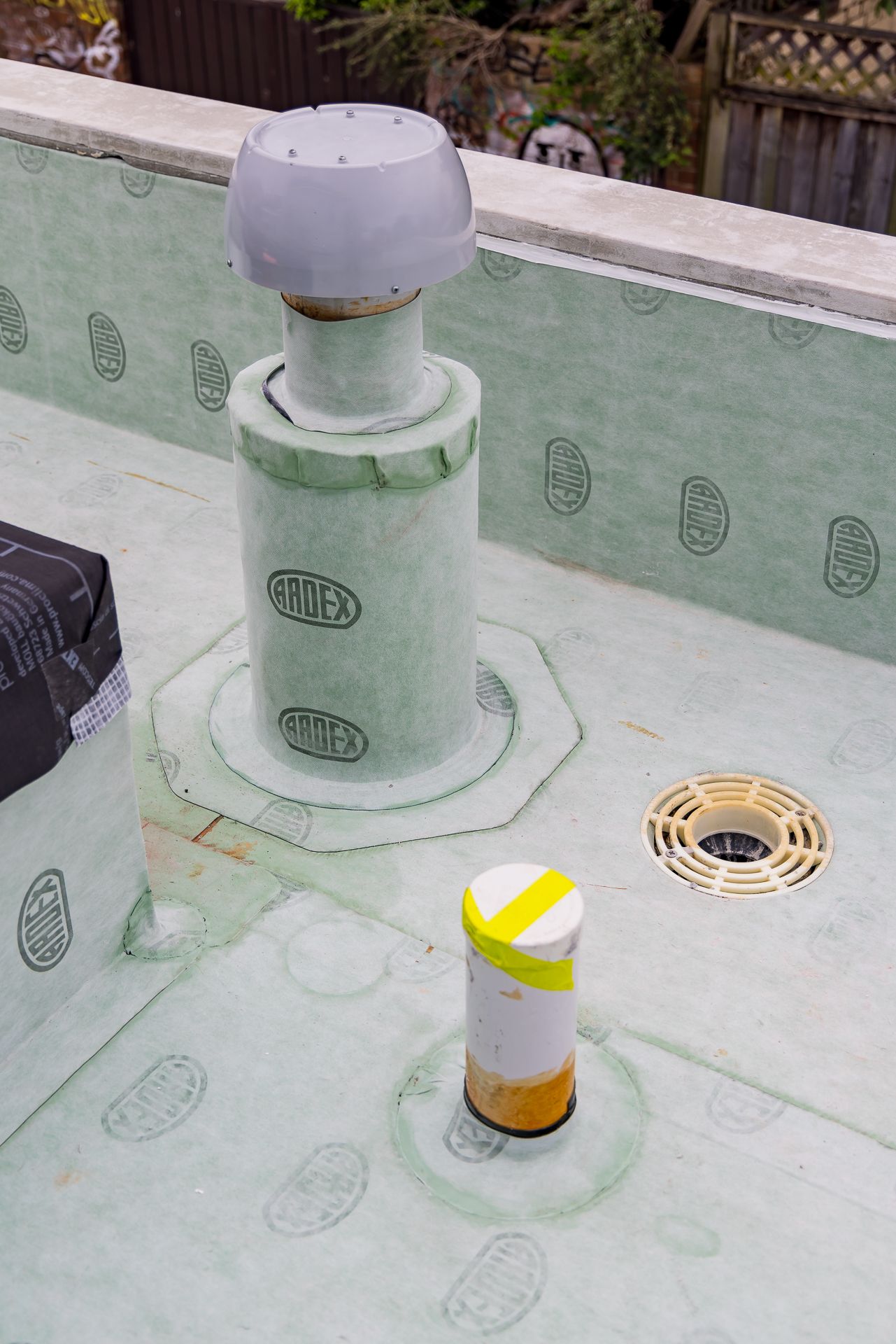 Rooftop waterproofing detail showing green membrane wrapped around a vent pipe, a floor drain and a taped marker on a construction site.