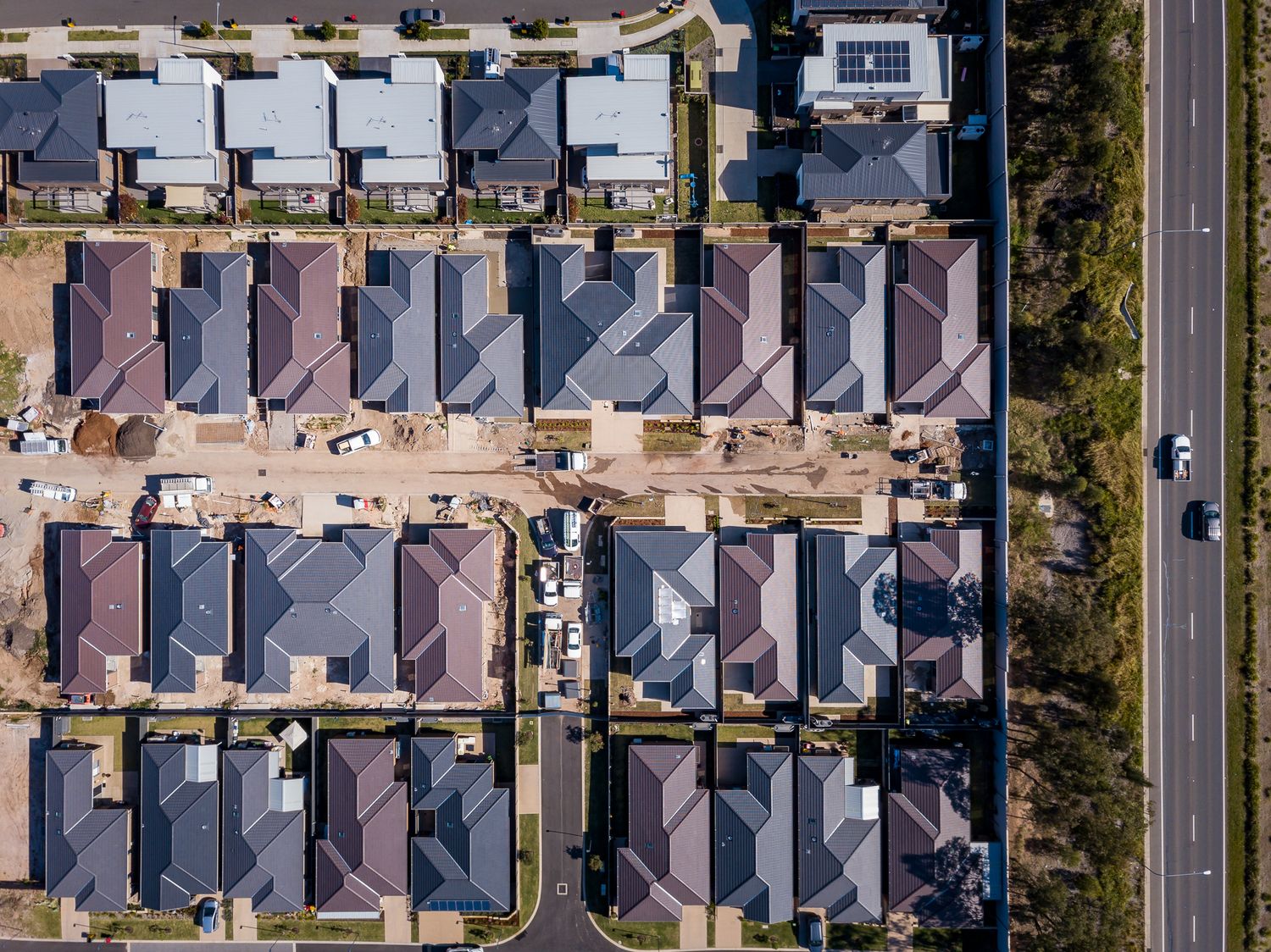 Top-down aerial view of a suburban street with new houses, grey and brown roofs, a building site and a busy main road alongside.