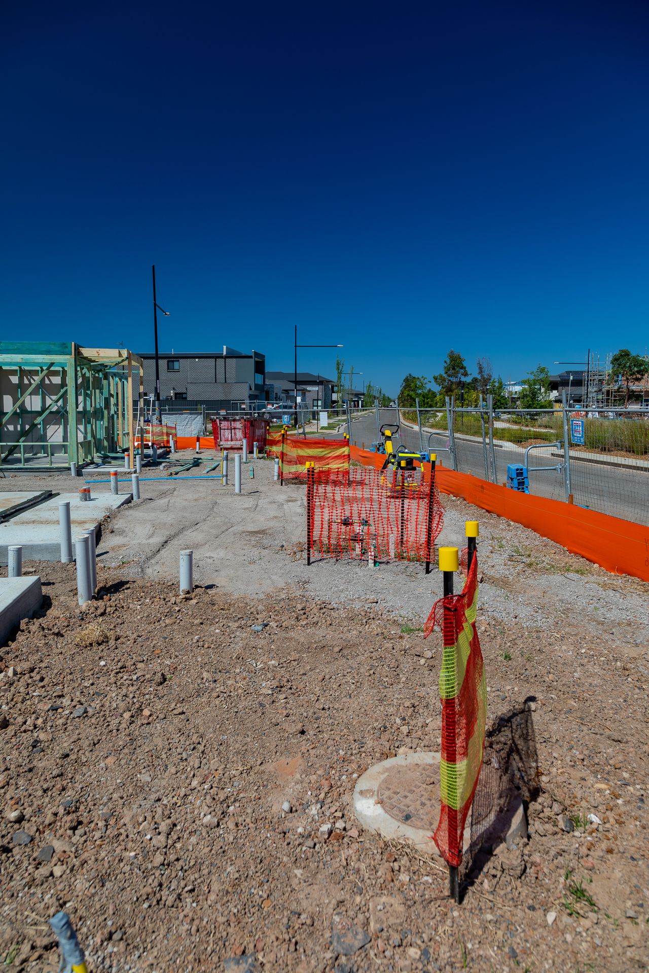 Construction site with orange safety fencing, plumbing pipes and a manhole cover on bare ground under a deep blue sky.