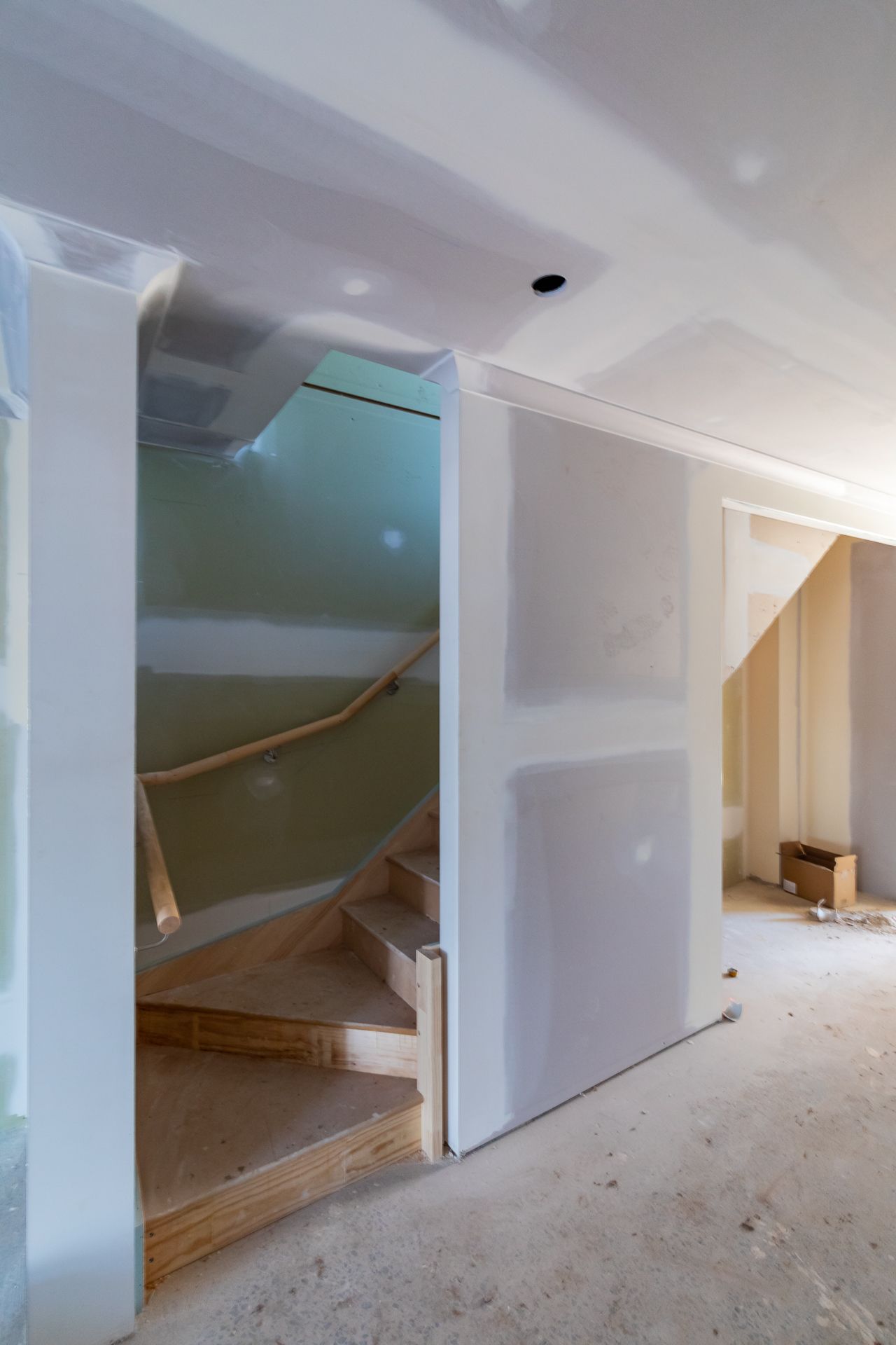 Interior of a home under construction with plasterboard walls, a timber staircase with handrail and a bare concrete floor.