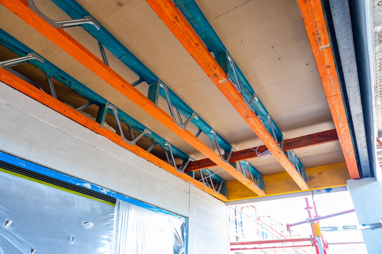 Underside of a building under construction showing orange and blue steel joists, ceiling panels and scaffolding outside.