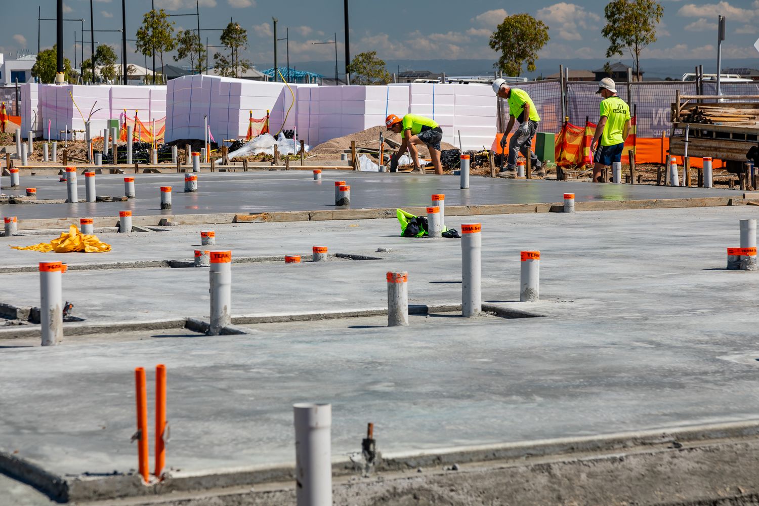 Two workers in yellow hi-vis shirts prepare a concrete slab with orange-capped pipes and wrapped materials in the background.