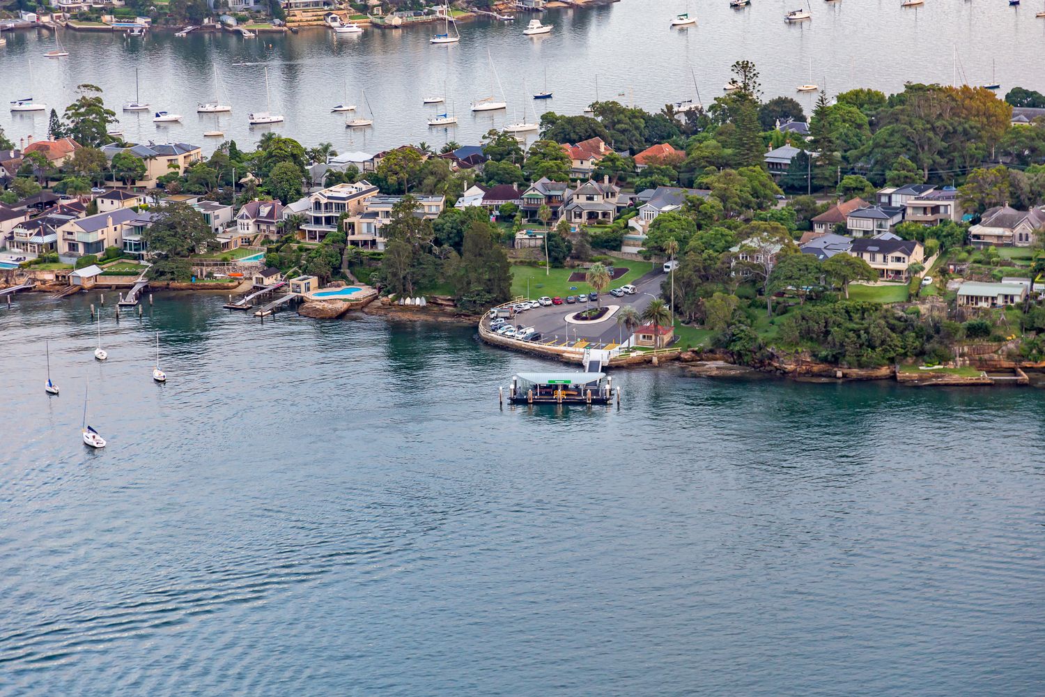 Aerial view of a small ferry wharf on a harbour foreshore surrounded by waterfront homes, green trees and moored boats.