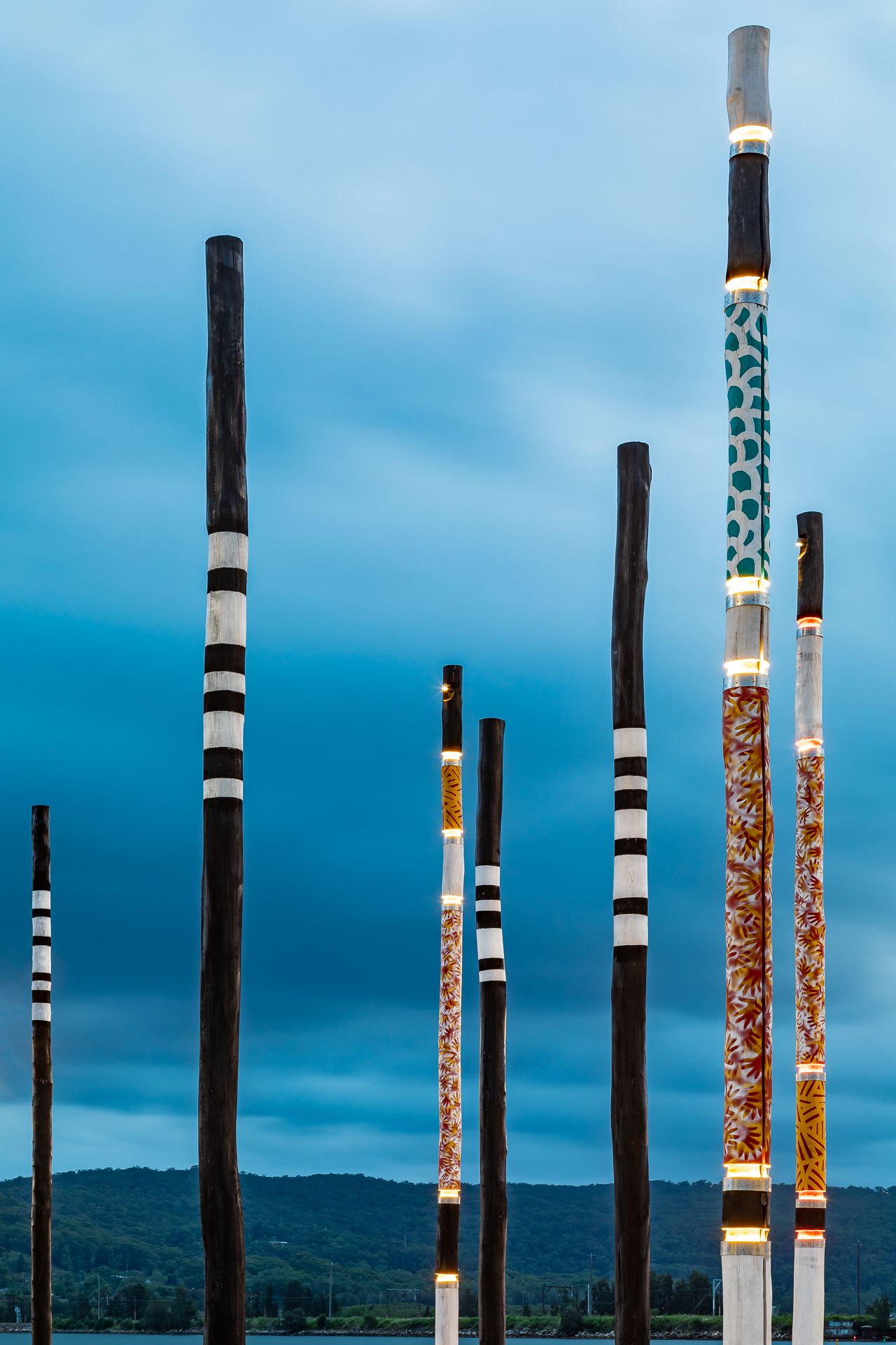 Tall decorated poles with black and white bands and orange and teal Indigenous-style patterns rise against a moody blue cloudy sky.