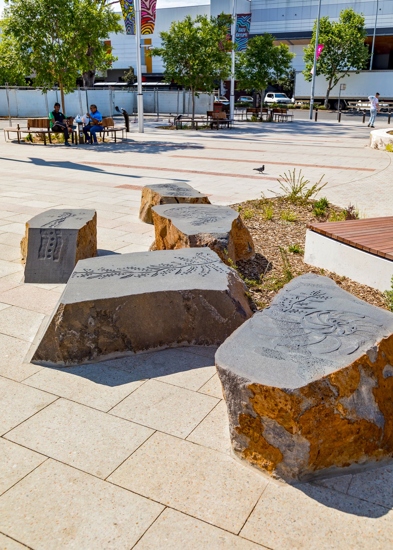 Carved sandstone boulder seats with engraved patterns in a paved public plaza, with people sitting on benches in the background.