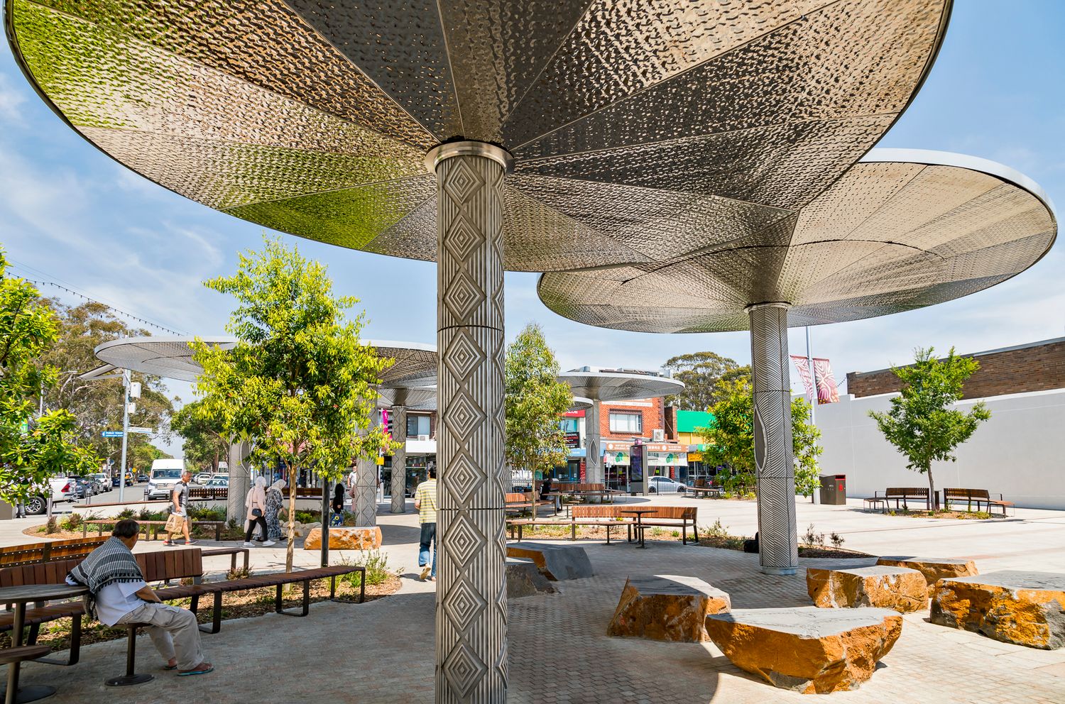 Perforated metal canopy shade structures above a sunny public square with people seated on benches and sculpted boulder seating.