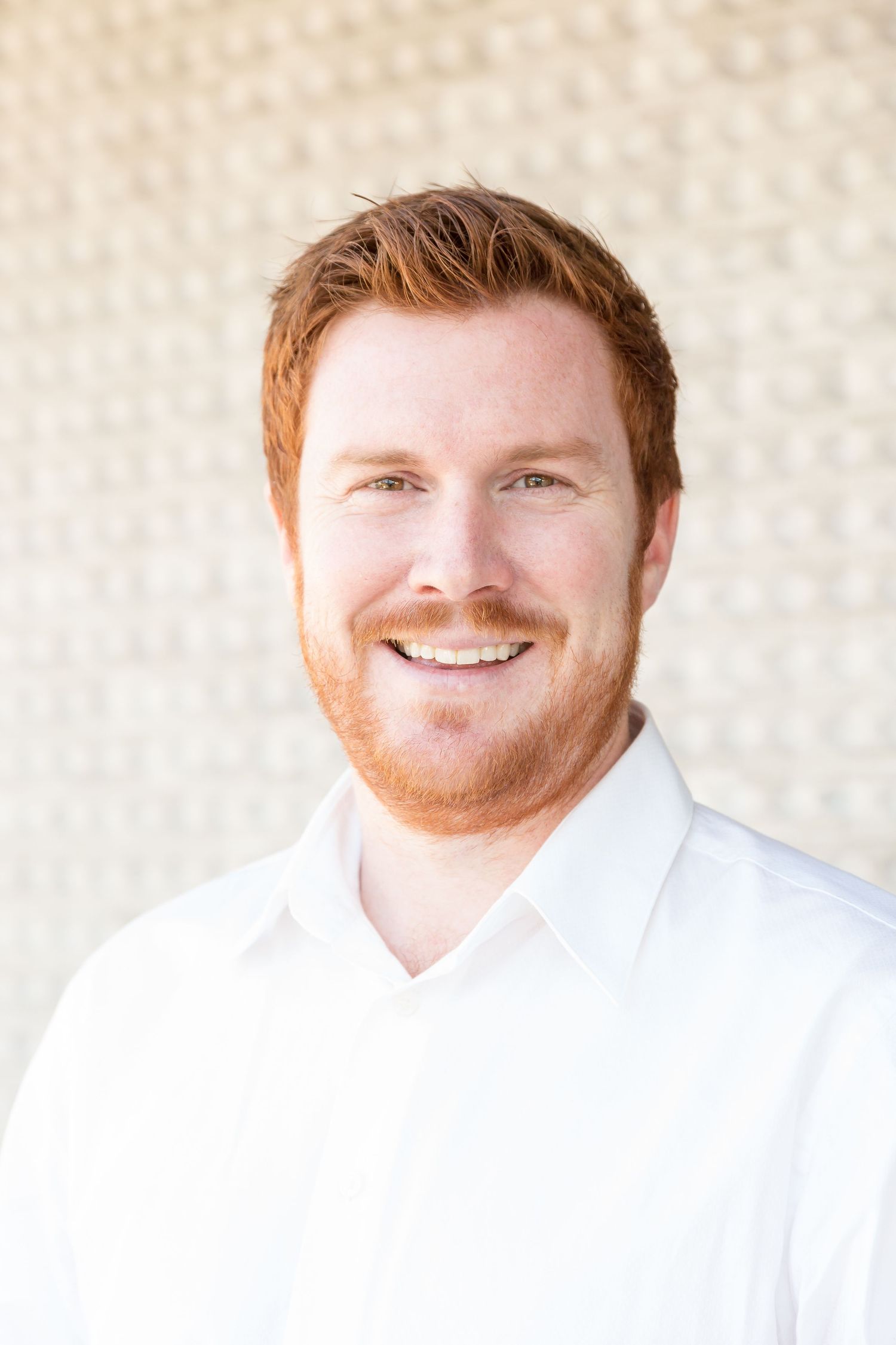 Smiling red-haired man with a beard wears a white collared shirt in a bright professional headshot against a textured light wall.