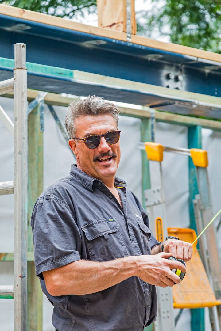A smiling mustached man in sunglasses and a gray work shirt holds a tape measure at a construction site with scaffolding.