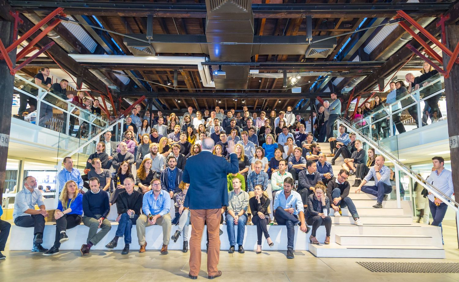 Large group of people gathered on a staircase and balconies inside a bright industrial-style office while a man addresses them.
