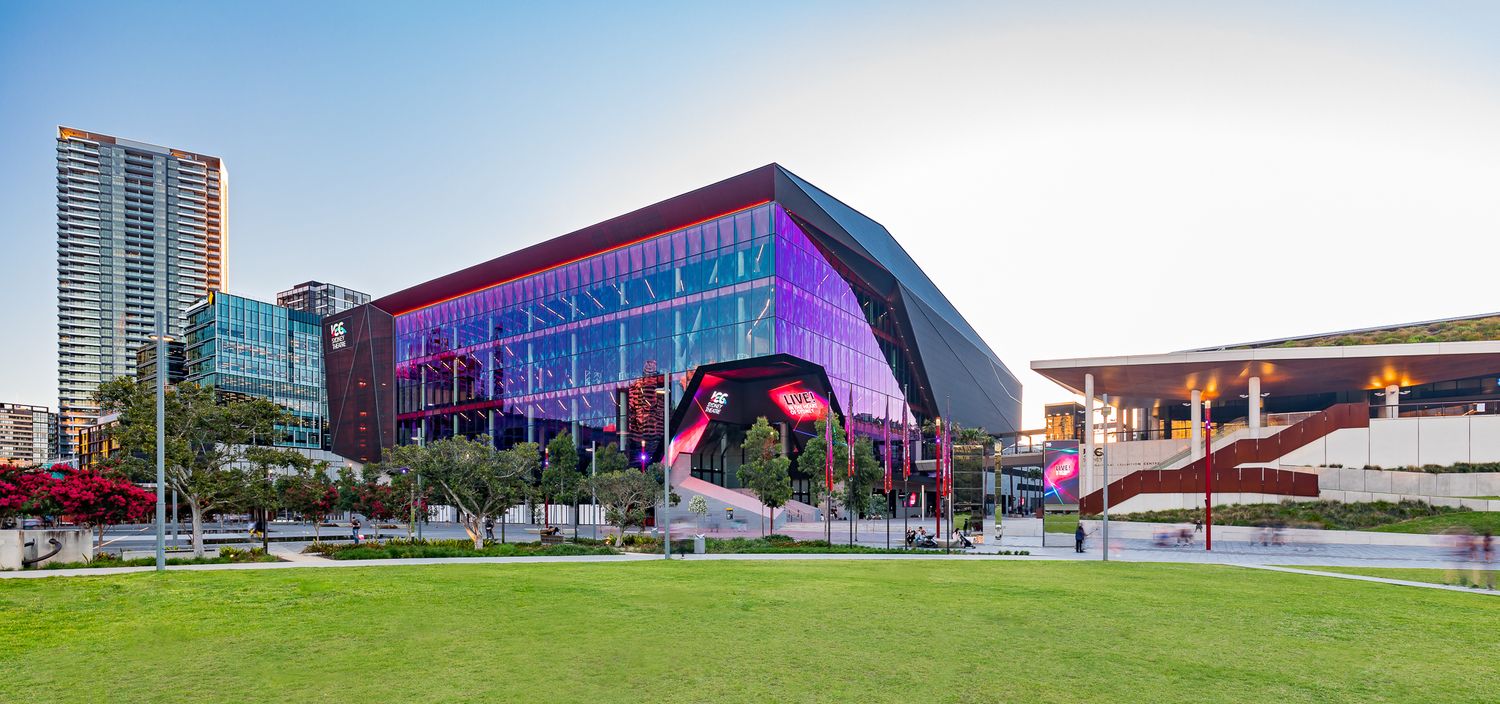Sleek angular convention centre with purple-tinted glass facade viewed across a green lawn on a bright sunny afternoon.