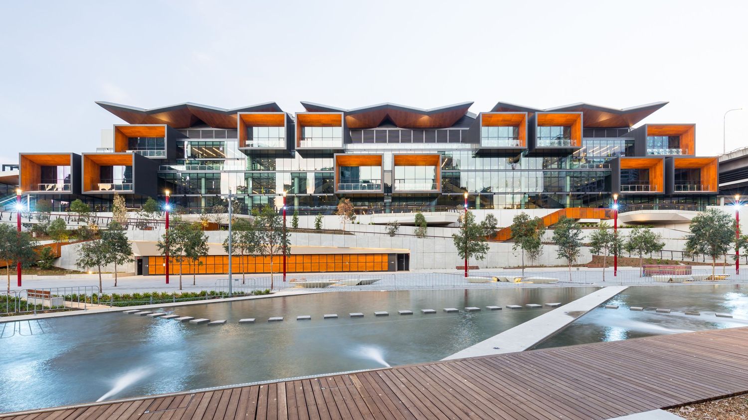 Modern terraced building with orange timber-framed balconies overlooking a reflective water feature and wooden boardwalk.