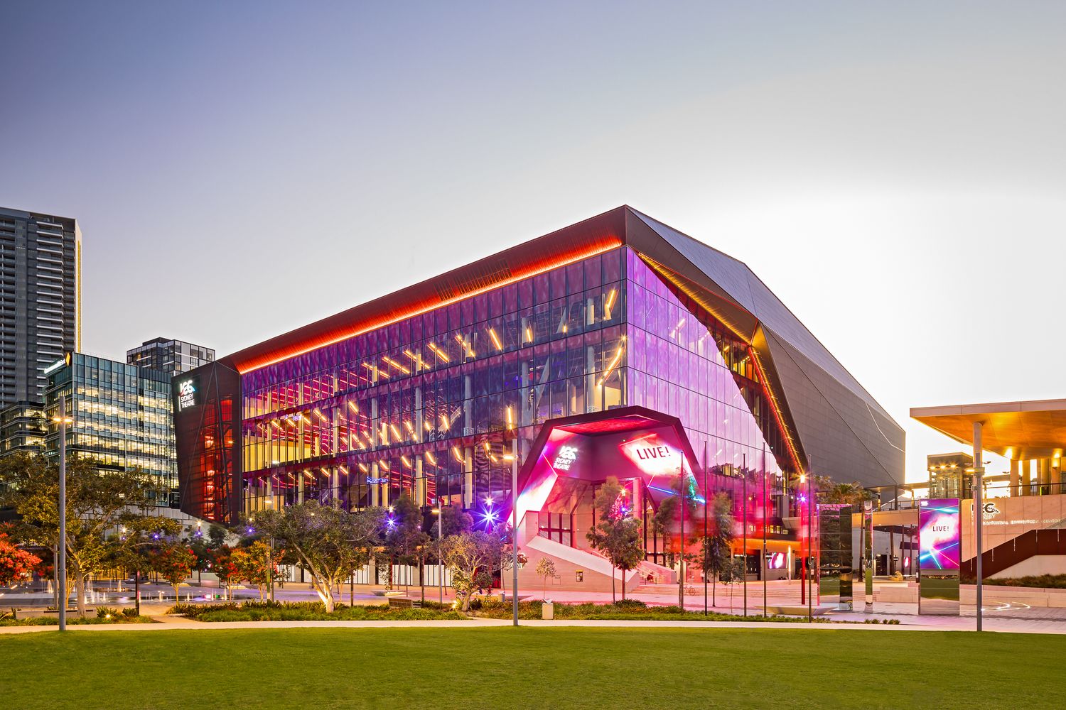 Glass convention centre glowing at twilight with red lighting, vibrant purple accents, a landscaped lawn and city towers in the background.