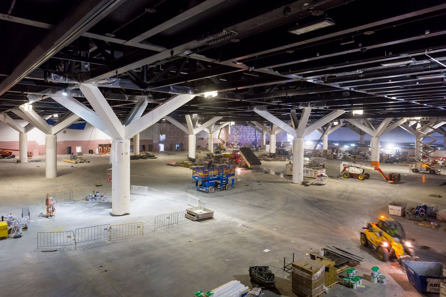 Large construction-phase hall with branching white concrete columns, scattered equipment and a grid of exposed ceiling beams.