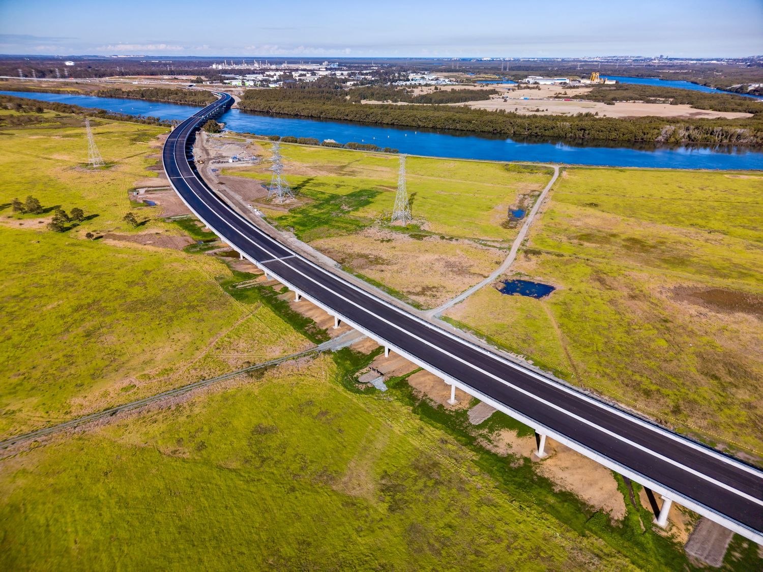 Aerial view of a curved elevated motorway bridge crossing green wetlands and grassland toward a wide river and distant industrial skyline.