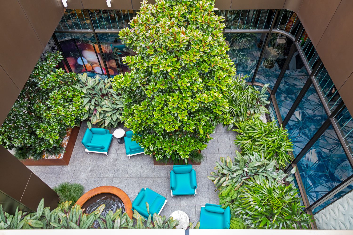 Aerial view of a courtyard with lush green plants, turquoise chairs and a colorful mural surrounded by glass-walled buildings.