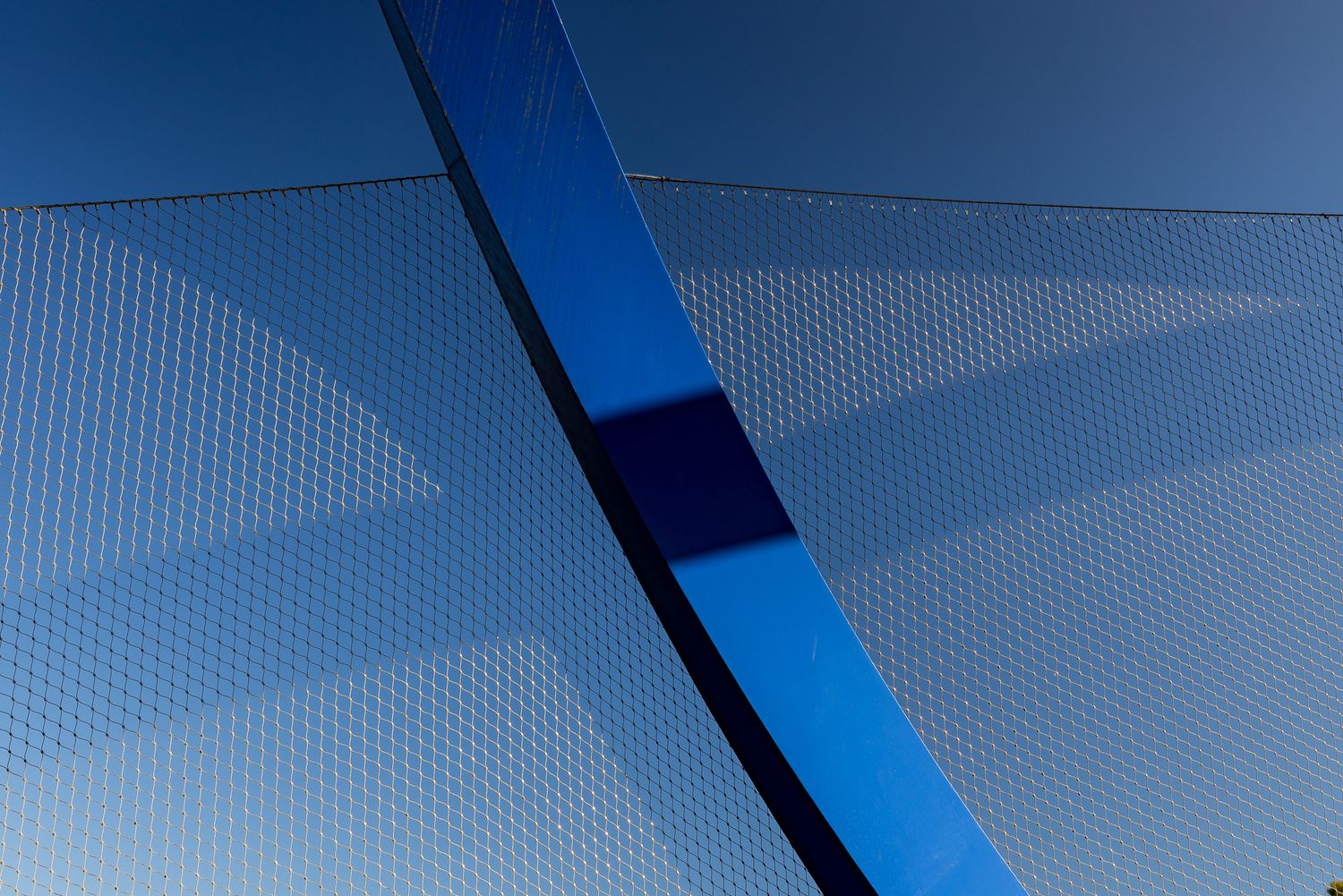 Abstract close-up of a curved blue steel beam against a fine mesh netting and clear blue sky.
