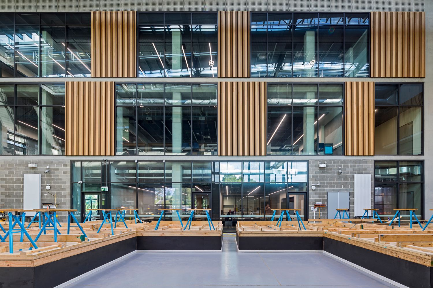 TAFE carpentry workshop with rows of workbenches and blue trestles in front of a timber-clad, glass-fronted multi-storey facade.