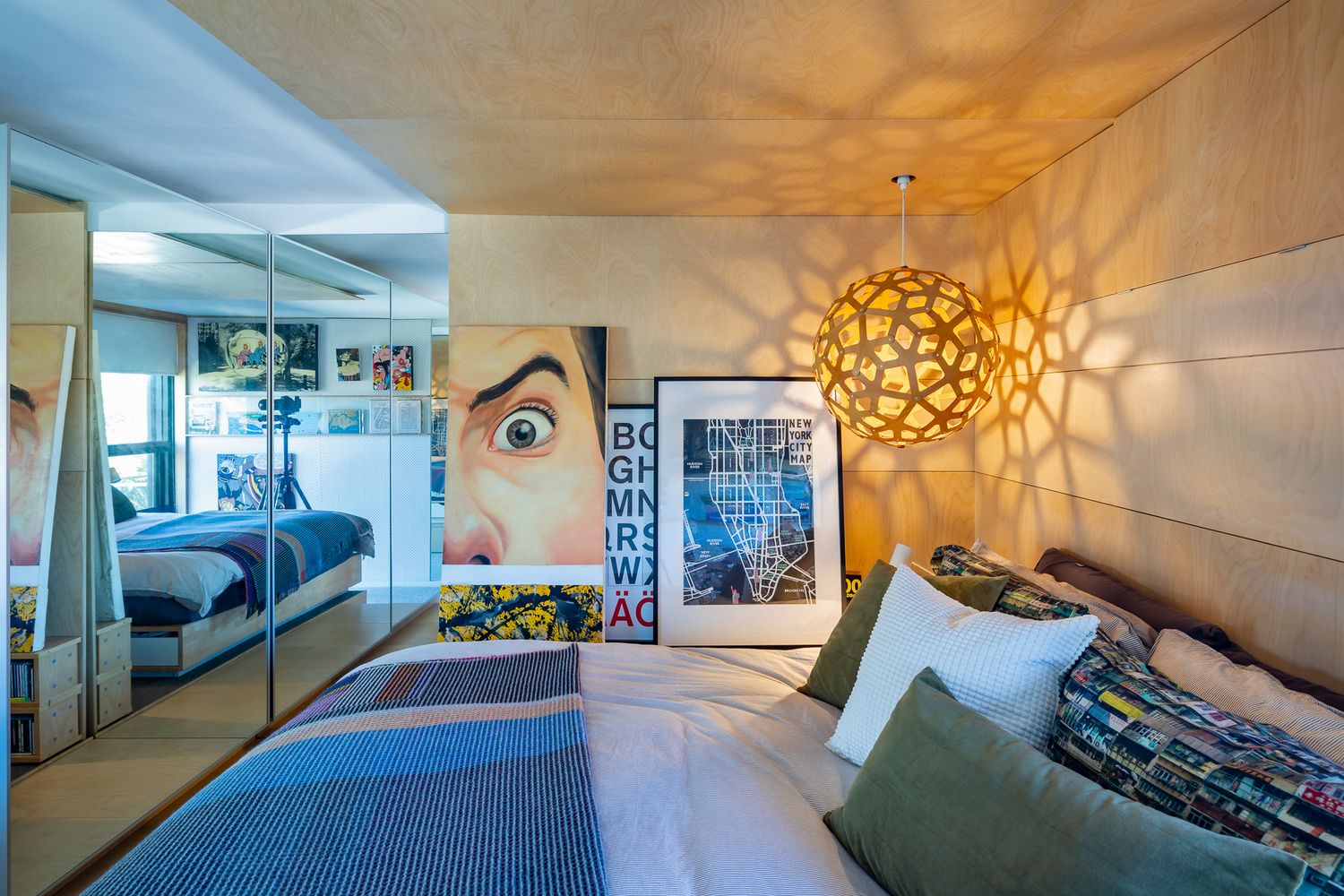 Cozy bedroom with a patterned throw, colorful cushions, pop-art eye poster, and a geometric pendant light casting shadows.