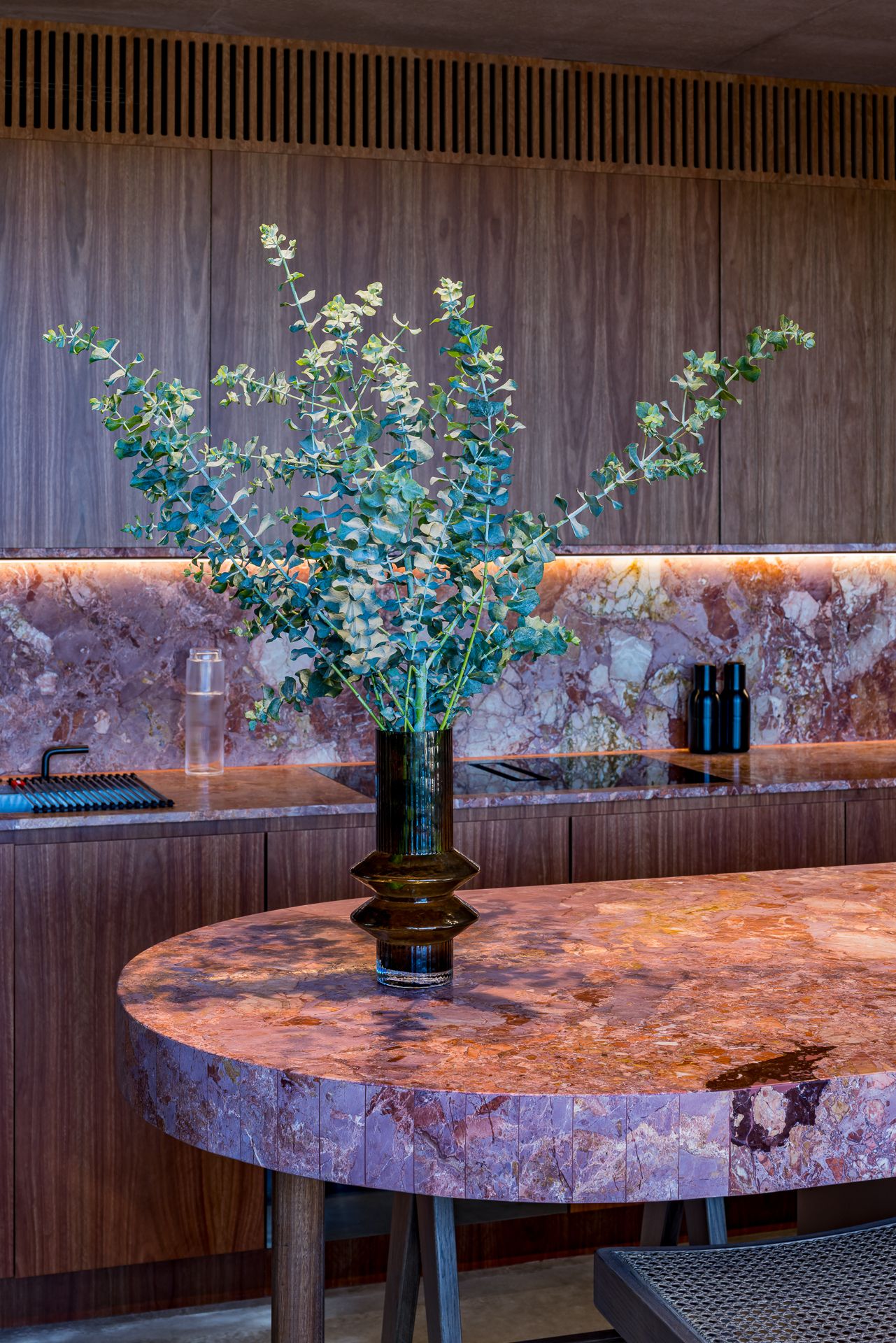 Eucalyptus branches in a dark glass vase on a pink marble oval table beside a walnut kitchen with matching marble backsplash.