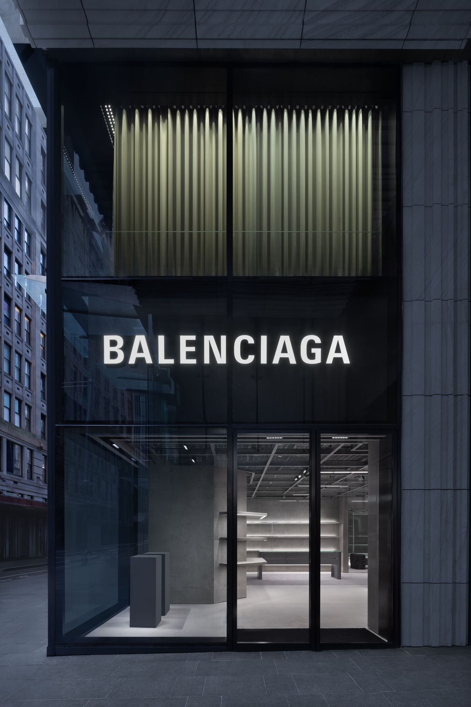 Two-story Balenciaga flagship storefront with a white illuminated sign, olive pleated drapery and a sleek modern glass entrance.