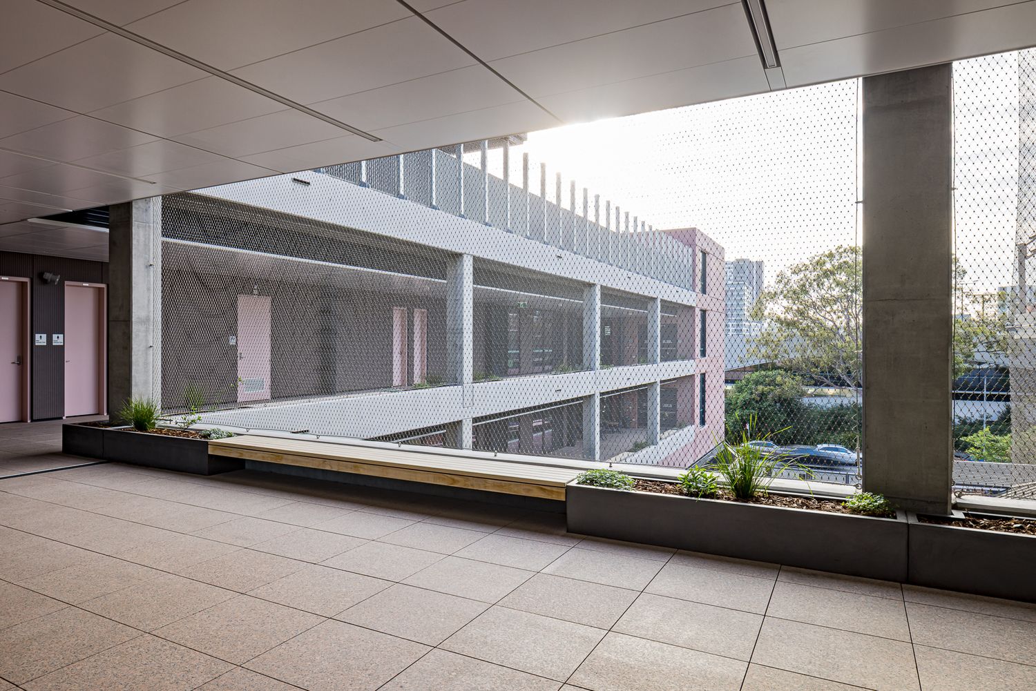 Spacious covered concrete terrace with planter boxes and a wooden bench overlooking a multi-level building and trees through mesh screening.