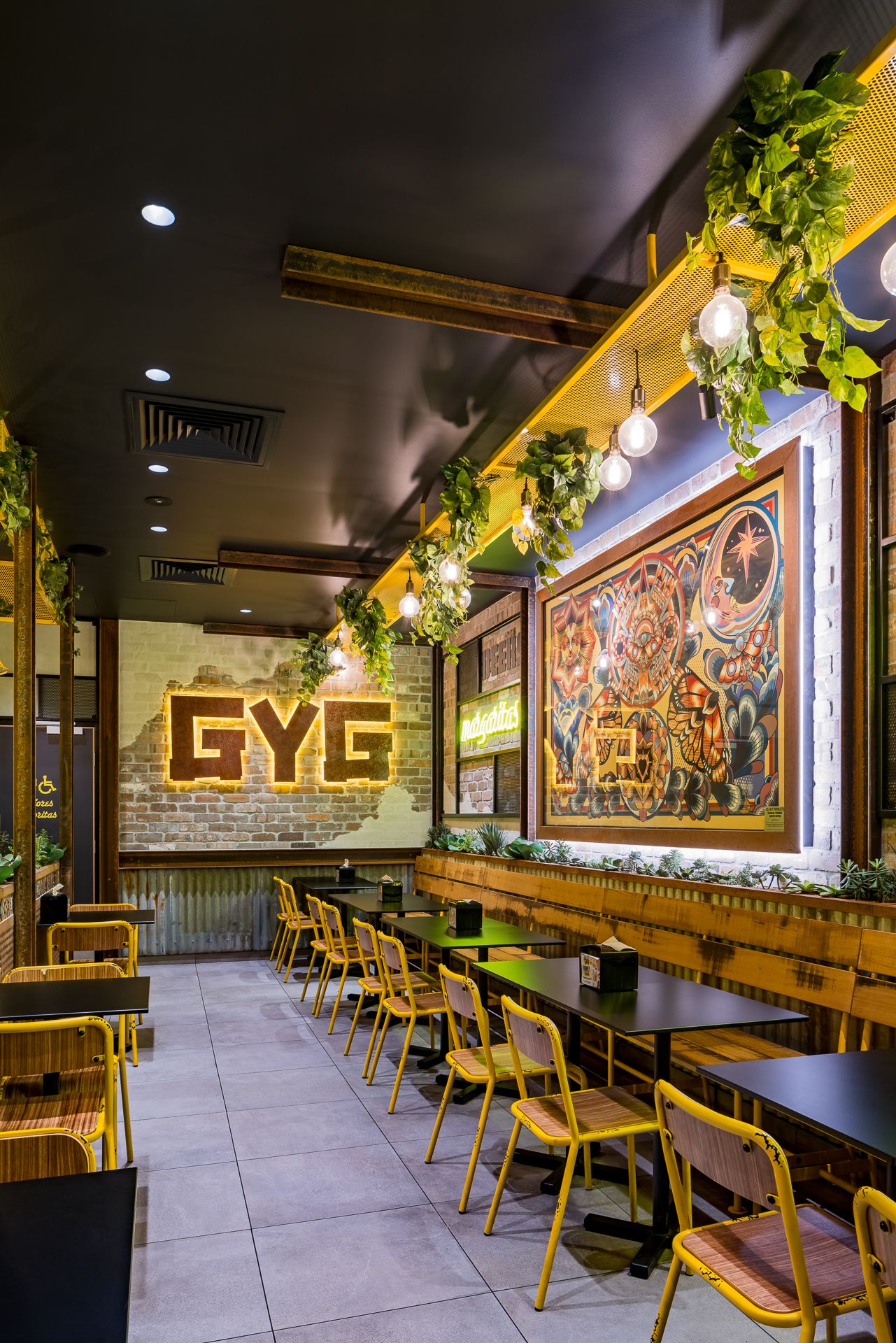 Colorful restaurant dining area with yellow chairs, dark tables, a GYG brick wall sign, hanging plants and a decorative mural.