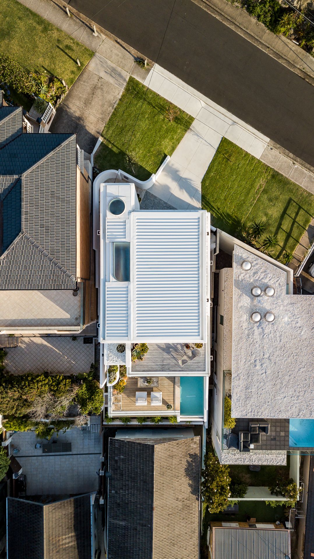 Overhead aerial view of a white contemporary home with a pool amid neighbouring rooftops and lawns.