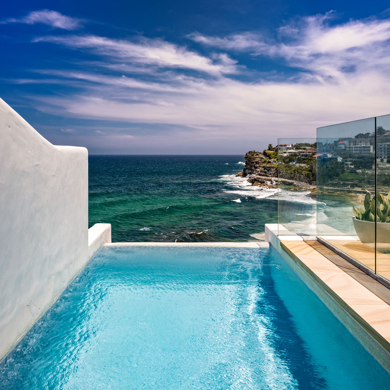Infinity-edge pool with glass balustrade overlooking turquoise ocean waves and a rocky headland under a dramatic blue sky.