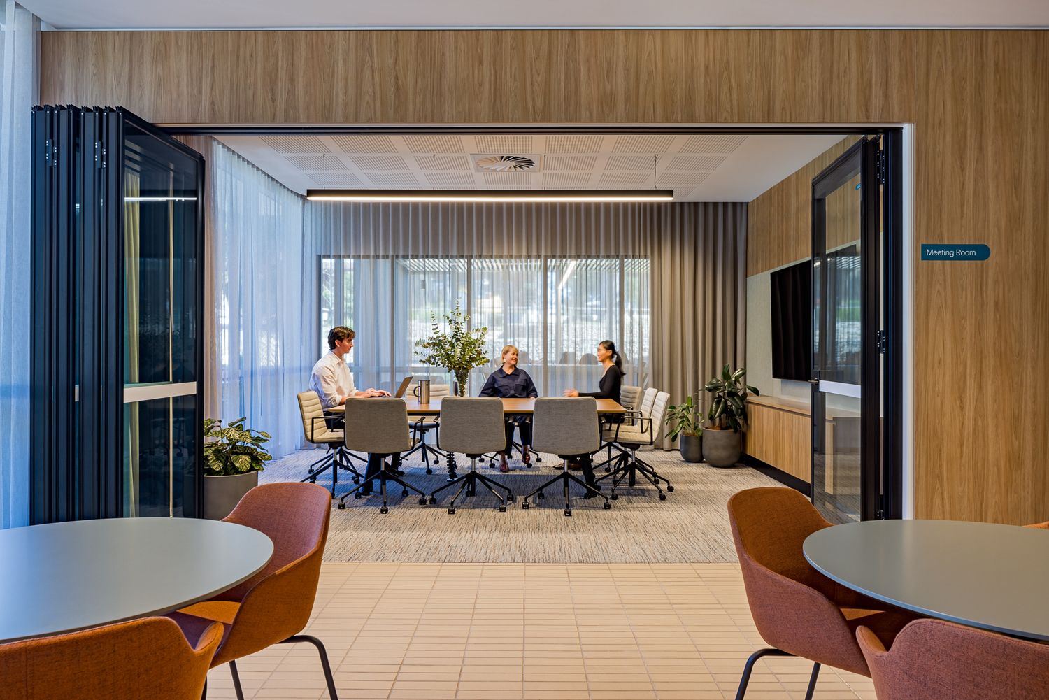 Three colleagues meet around a conference table in a glass-walled modern office meeting room with wood paneling and orange chairs in the for
