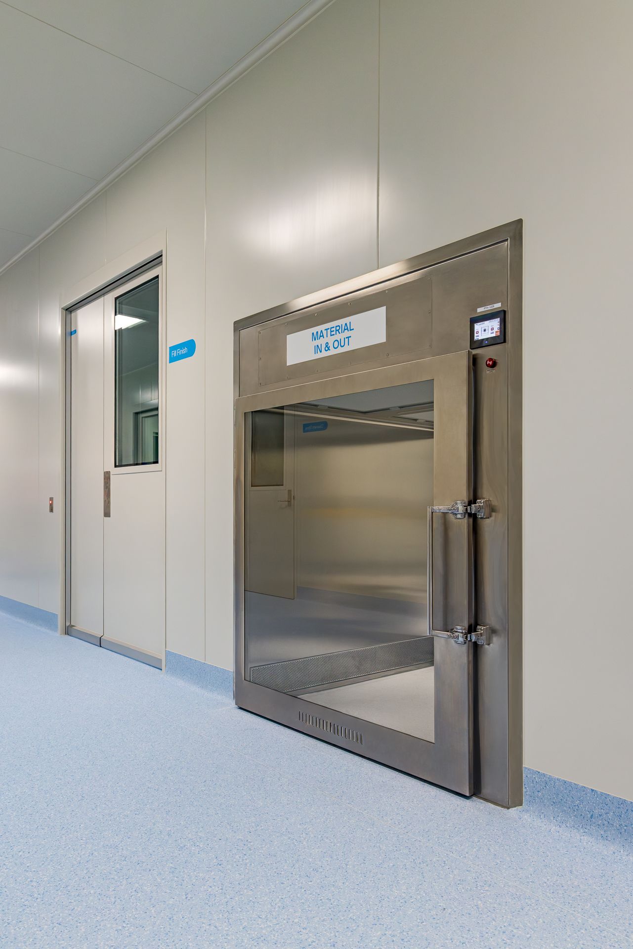 Stainless steel material airlock door labeled Material In and Out in a clean corridor of a pharmaceutical cleanroom.