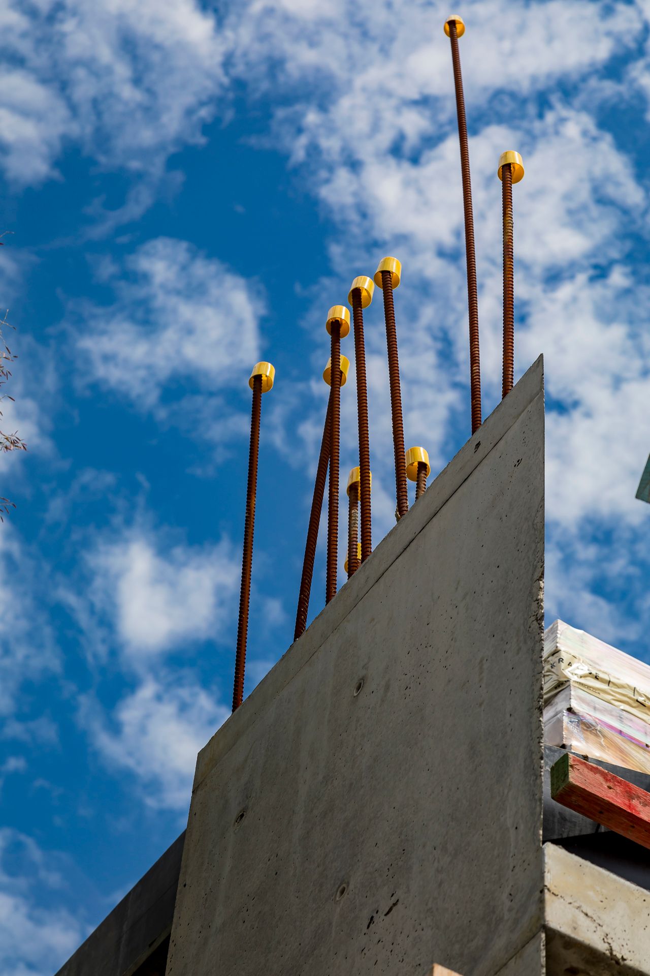 Rusted steel reinforcing bars with yellow safety caps rising above a concrete wall against a blue sky with white clouds.