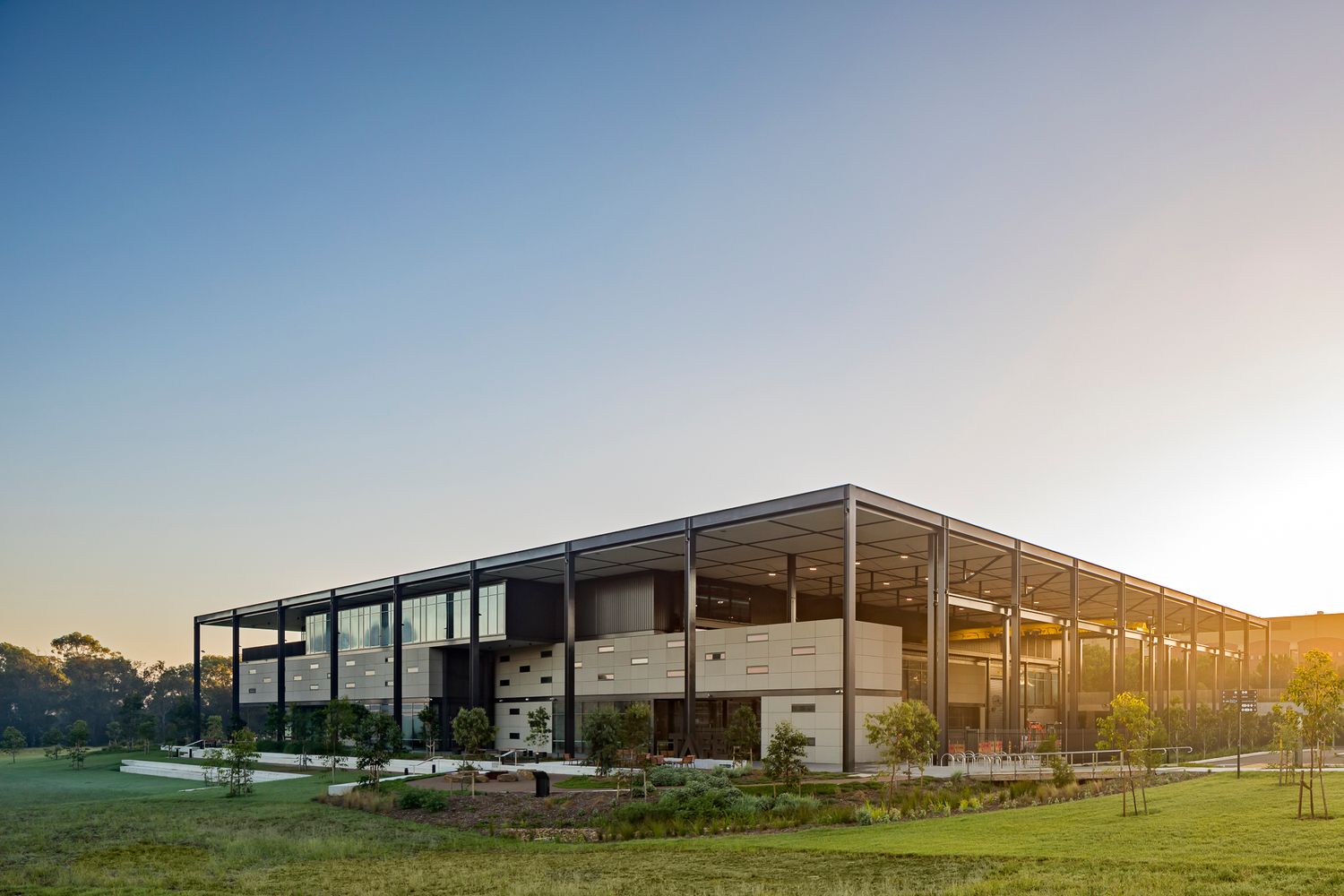 Contemporary steel-framed building with stone cladding and a broad roof canopy beside native landscaping in warm evening light.